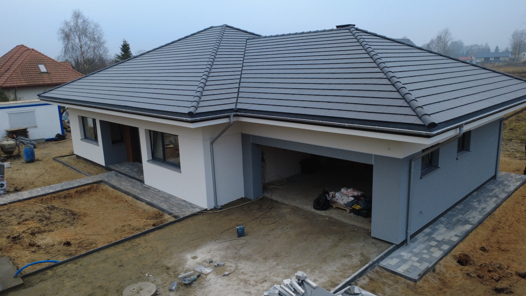 Nowoczesny dom jednorodzinny z szarą dachówką i elewacją w odcieniach bieli i szarości. Widoczne prace wykończeniowe wokół budynku, kostka brukowa i przygotowany teren pod ogród.