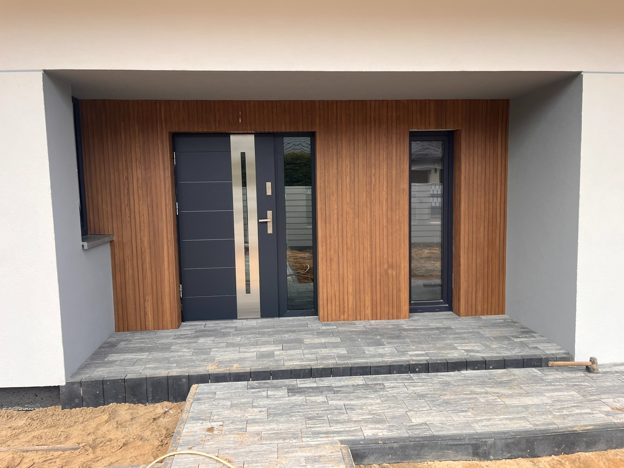 Nowoczesne wejście do domu z szarą stolarką drzwiową i okładziną ścienną imitującą drewno. Ułożona kostka brukowa na podeście. Widoczny młotek.