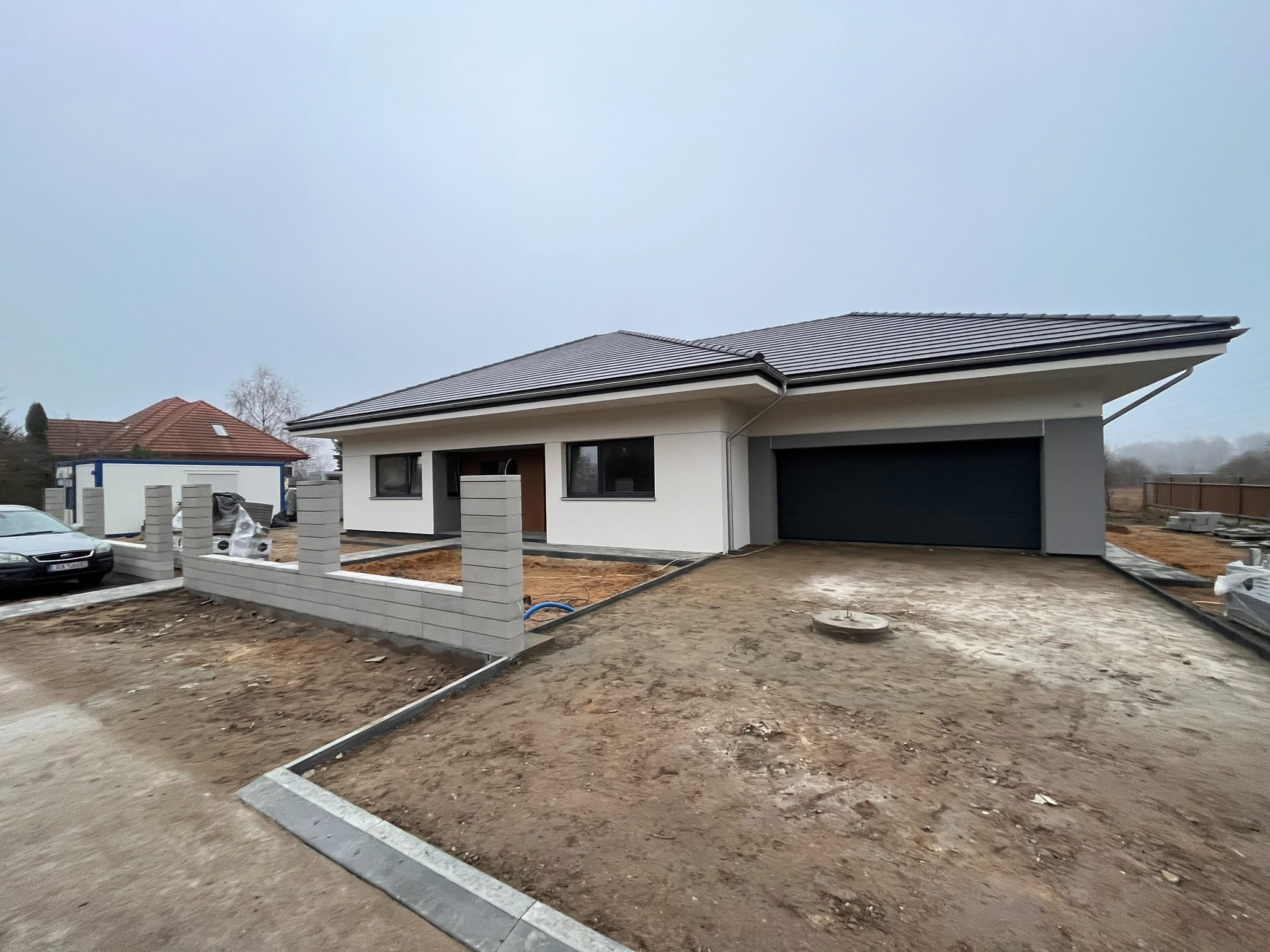 Nowoczesny dom jednorodzinny z garażem w trakcie wykończenia. Widoczny fragment ogrodzenia z szarej kostki, podjazd i elewacja w jasnych barwach. Pochmurne niebo w tle.