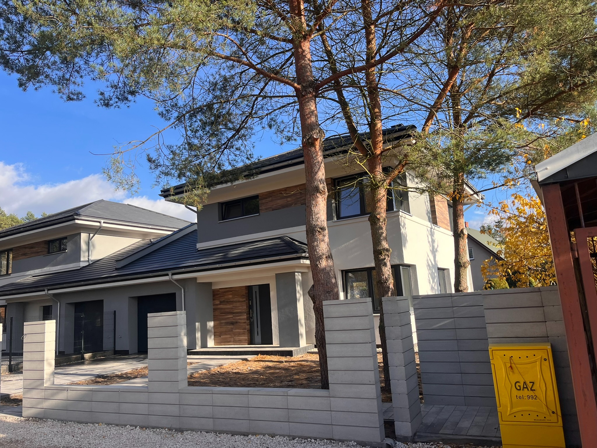 Nowoczesny dom jednorodzinny z szarą elewacją i drewnianymi akcentami, otoczony drzewami i betonowym ogrodzeniem. Widoczna skrzynka gazowa.