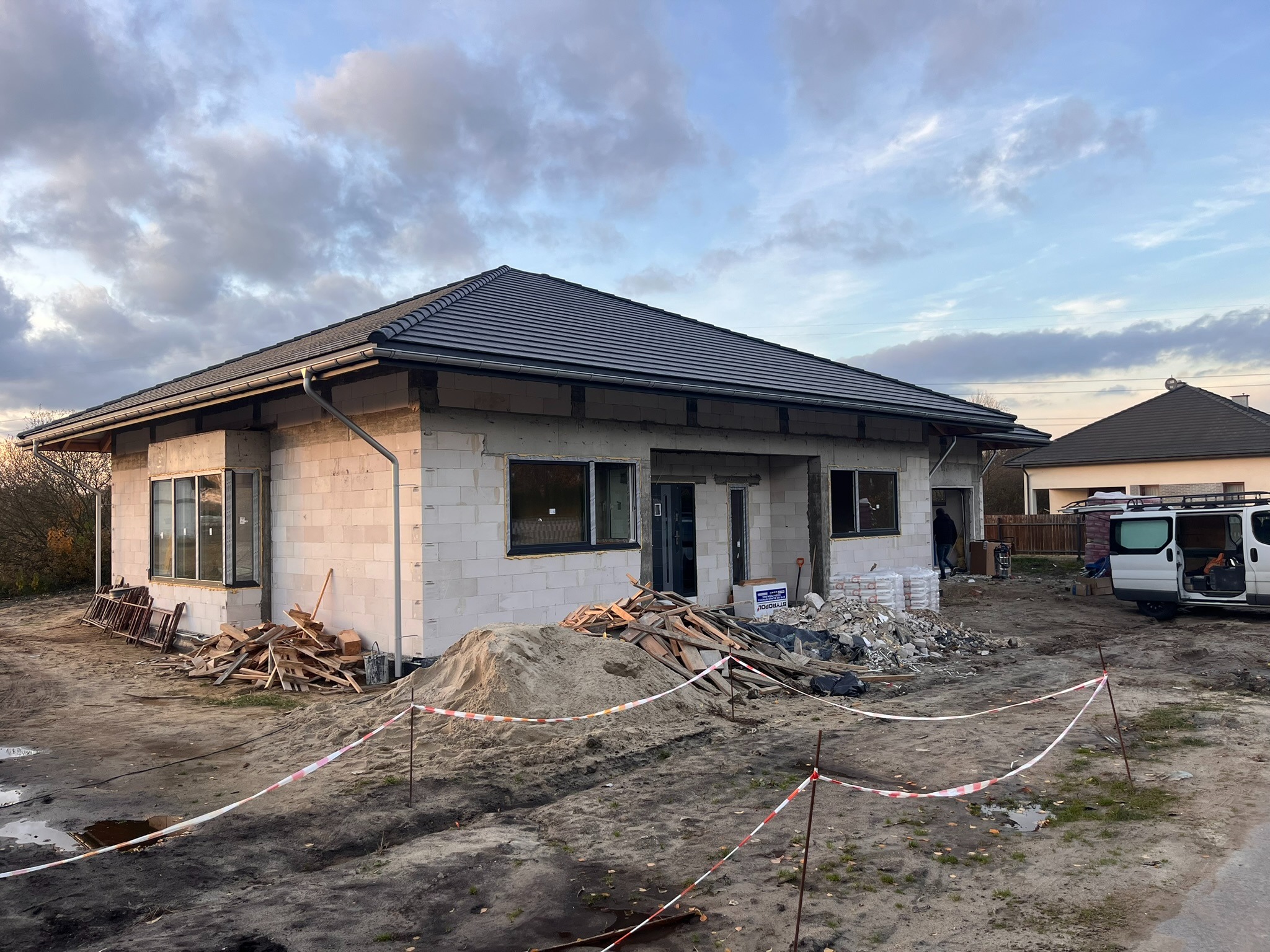 Surowy, parterowy dom z szarego bloczka, z ciemną dachówką i czarnymi oknami, w trakcie budowy. Na pierwszym planie piasek, gruz i taśma ostrzegawcza.