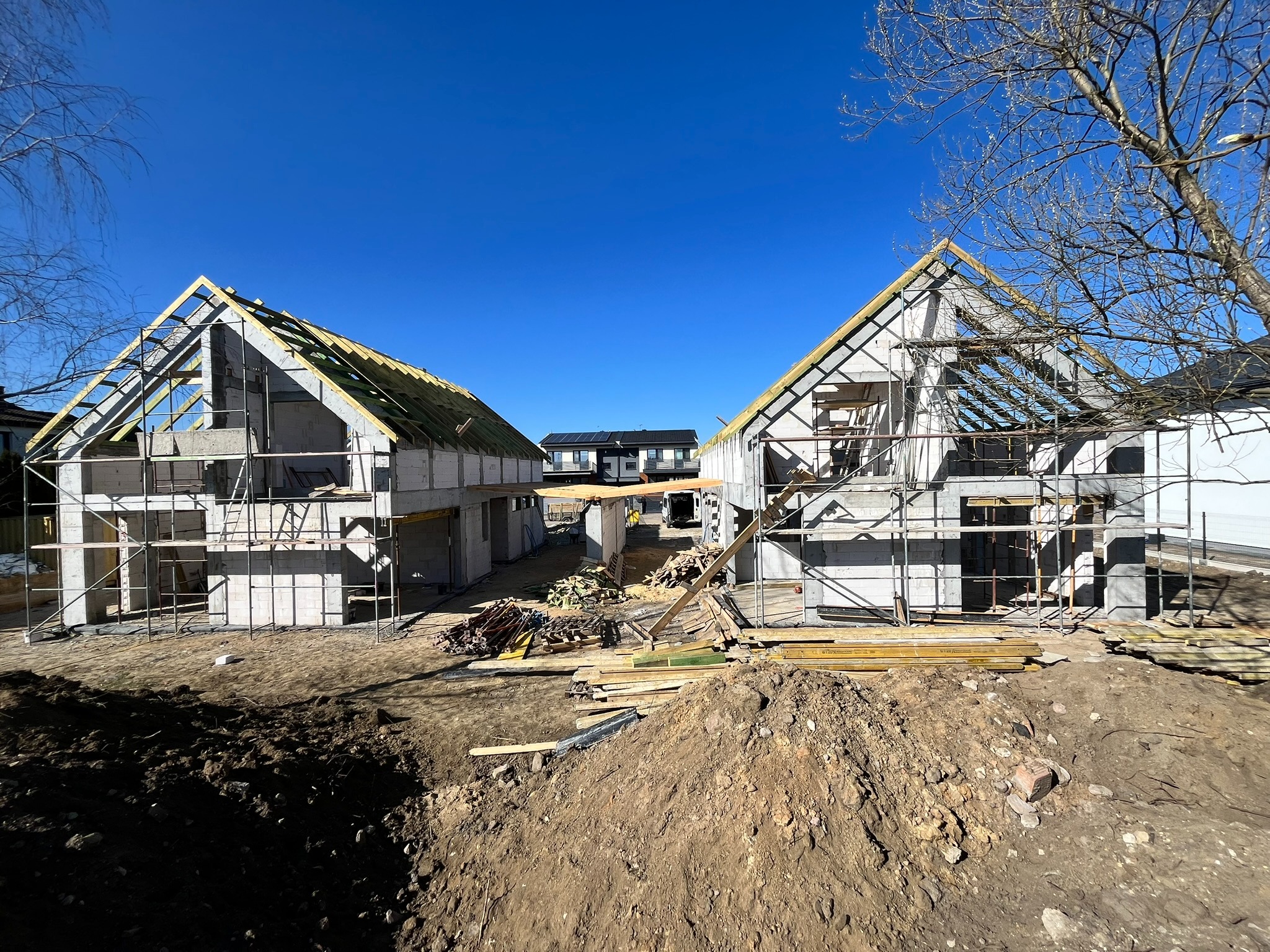 Budowa dwóch domów jednorodzinnych w stanie surowym otwartym, widoczne rusztowania i materiały budowlane. Błękitne niebo w tle.