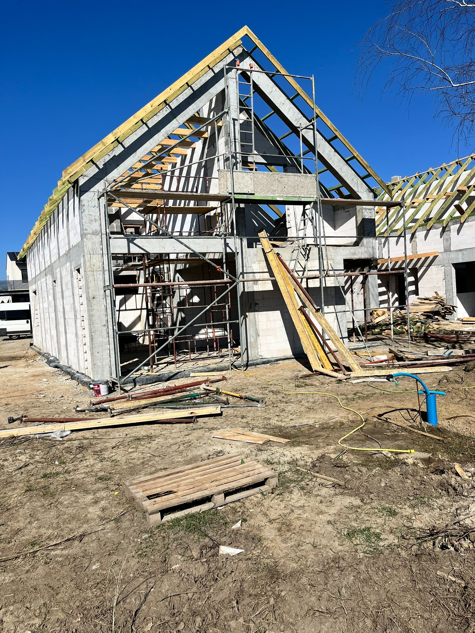 Surowy stan budynku w trakcie budowy: betonowe ściany, drewniana konstrukcja dachu, rusztowania. Widoczne materiały budowlane i narzędzia na placu budowy.