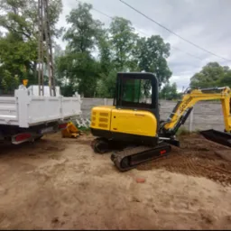 Żółta mini koparka GG3000 na gąsienicach obok białej wywrotki na placu budowy, widok z boku na tle betonowego ogrodzenia i drzew.