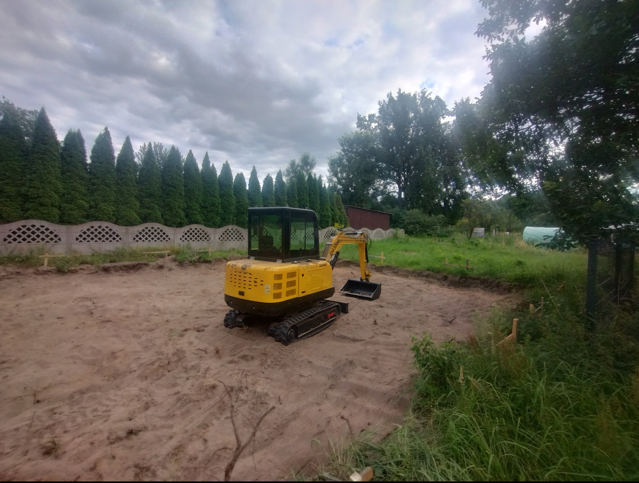 Żółta mini koparka na placu budowy, widoczne wykopane fundamenty i ogrodzenie z tujami w tle. Maszyna GG3000 gotowa do pracy.