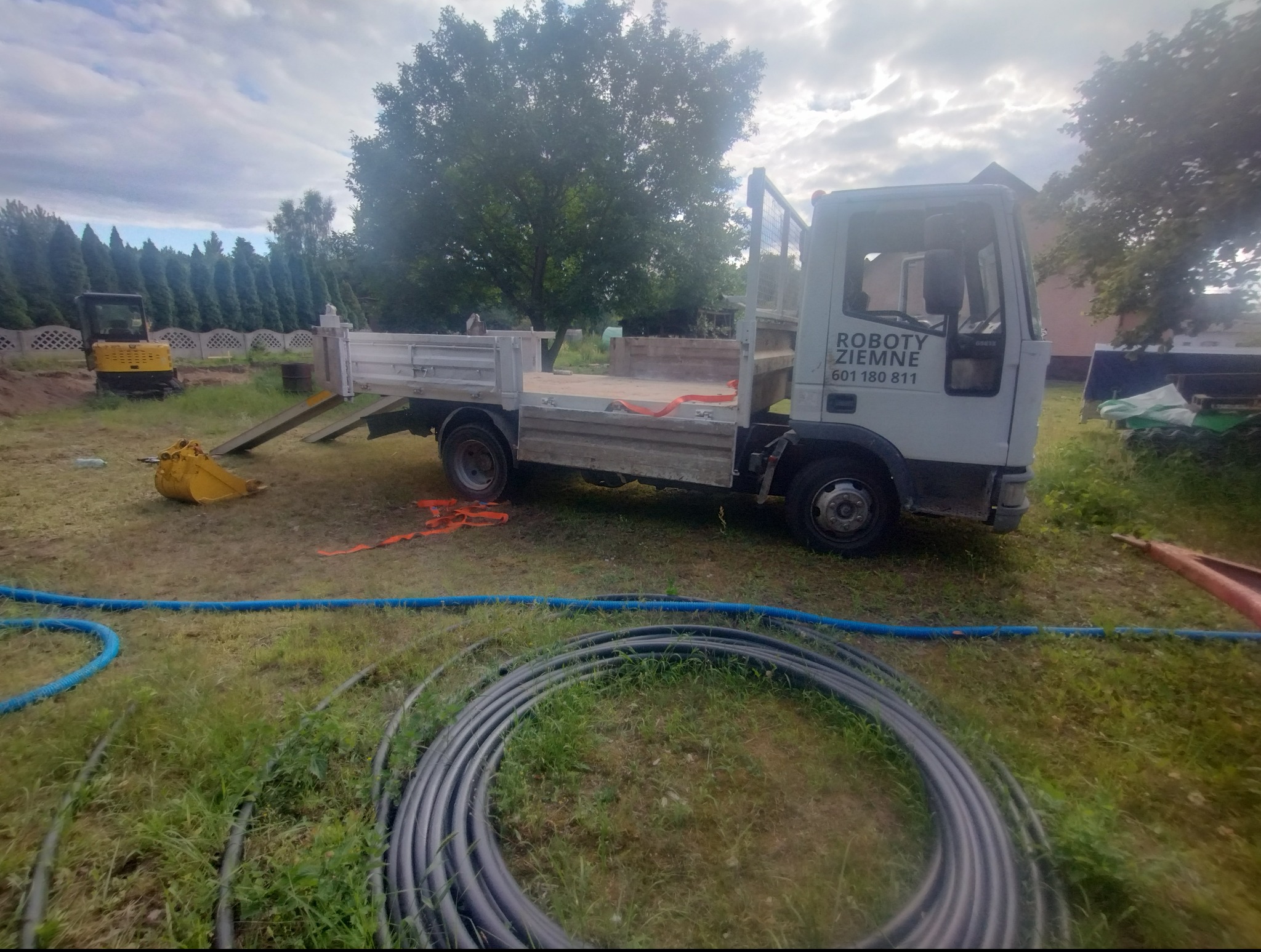 Ciężarówka z napisem 'Roboty Ziemne' na tle placu budowy z minikoparką i zwojami rur.