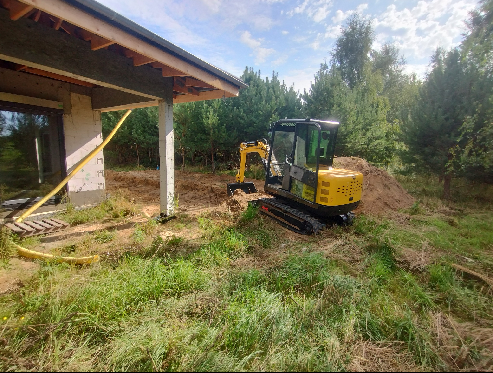 Żółta mini koparka przy fundamentach domu w trakcie prac ziemnych, widoczny wykop i hałda ziemi. W tle drzewa iglaste i błękitne niebo.