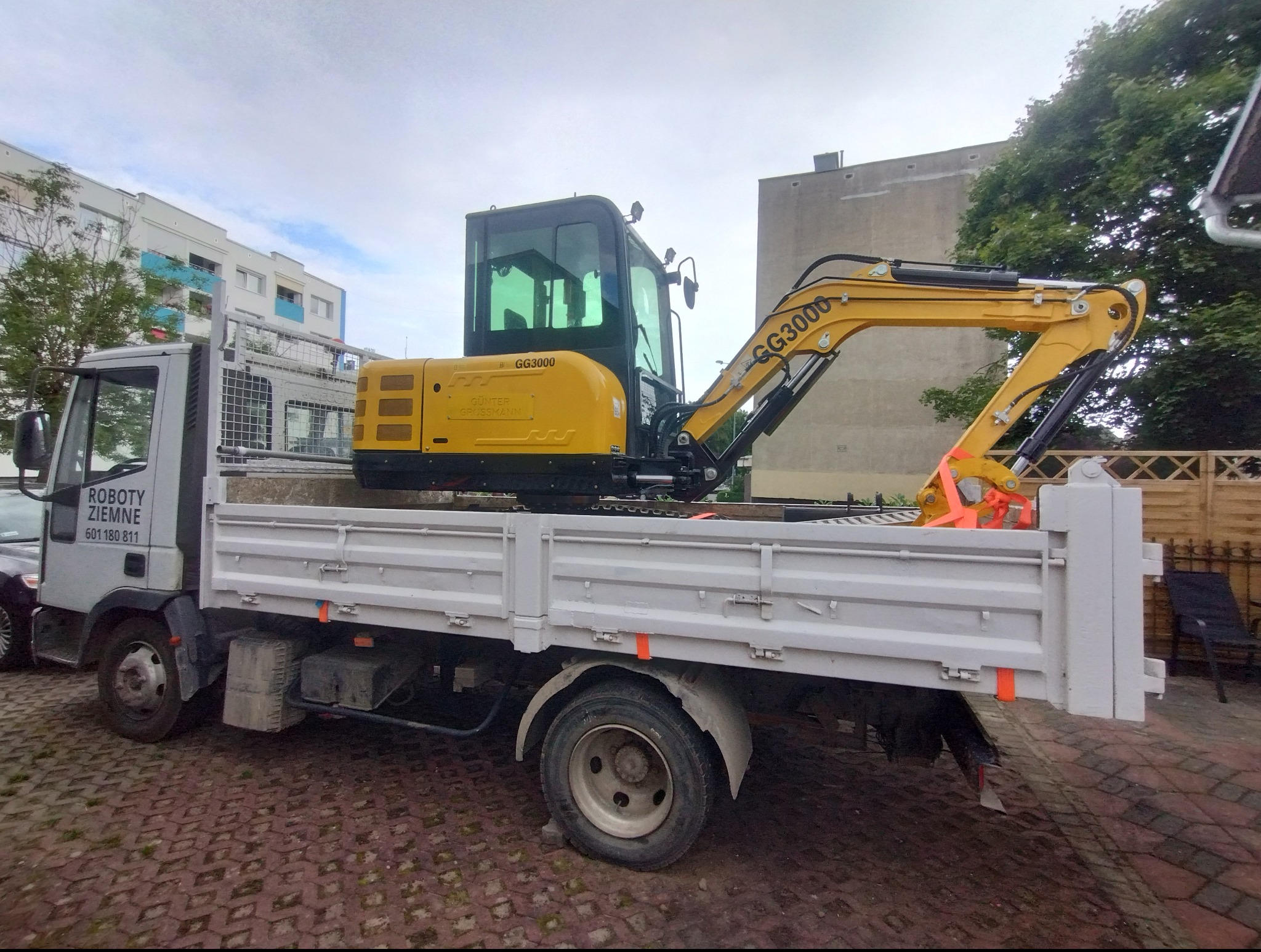 Żółta minikoparka GG3000 zabezpieczona pomarańczowymi pasami transportowymi na białej ciężarówce z napisem 'Roboty Ziemne' na tle budynków i drzew.