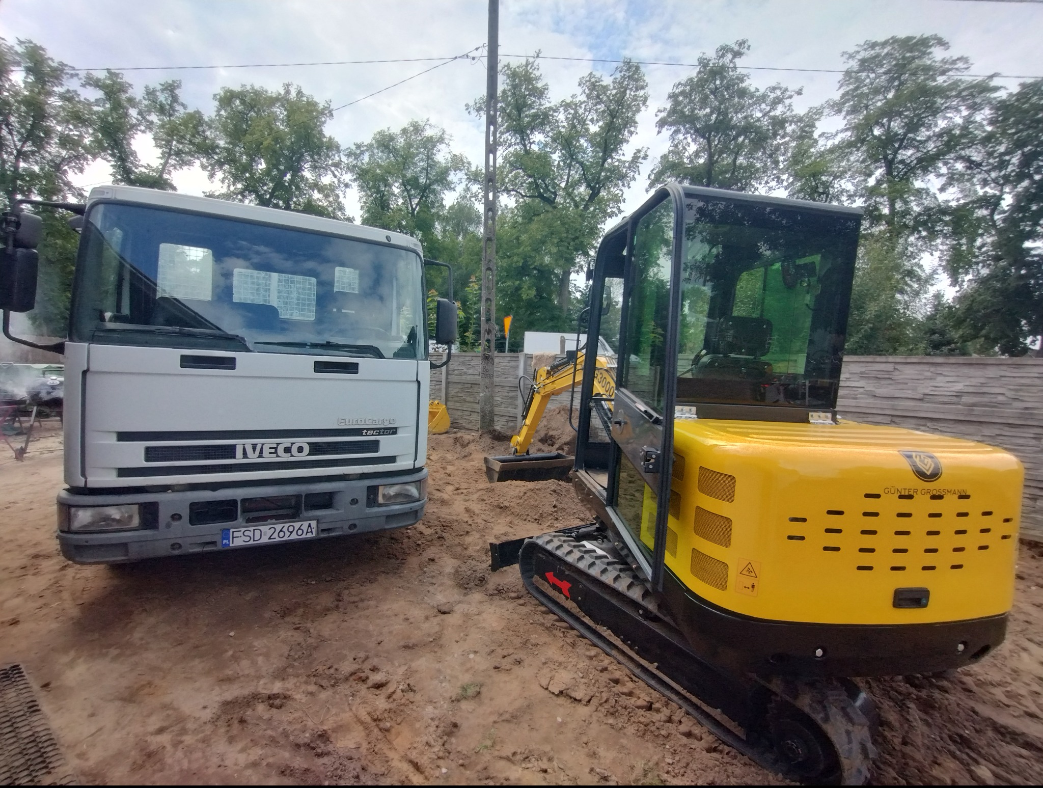 Żółta mini koparka Günter Grossmann i biała ciężarówka Iveco na placu budowy, ziemia, częściowo widoczny wykop. Sprzęt budowlany w trakcie pracy.