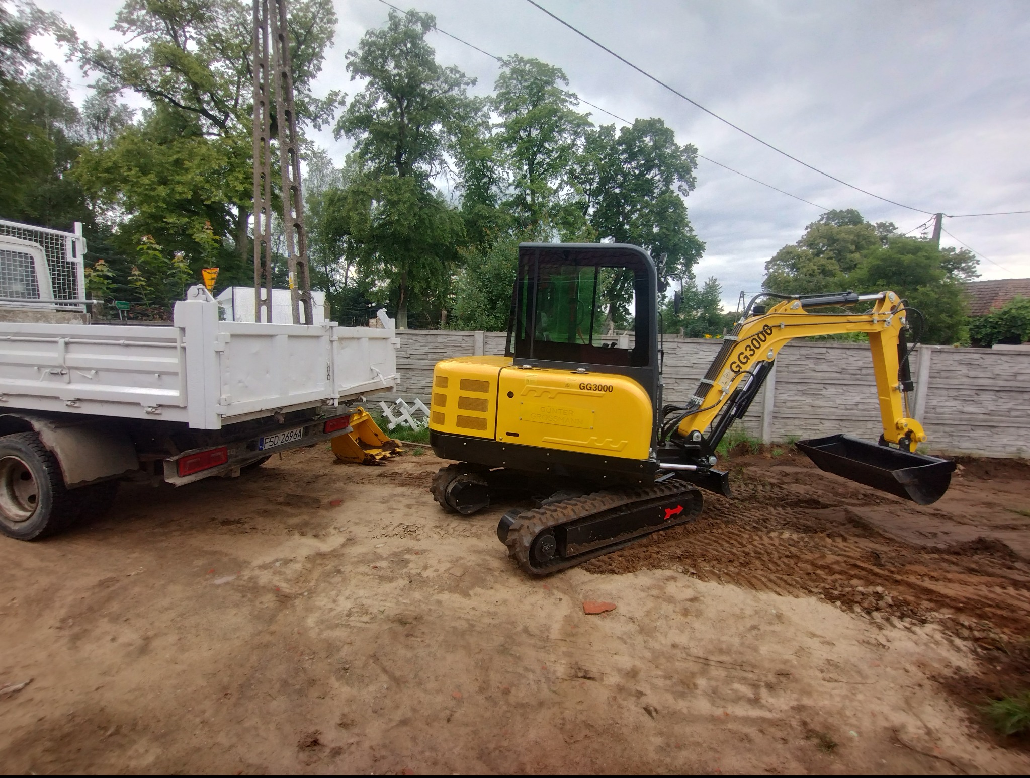 Żółta mini koparka GG3000 na gąsienicach obok białej wywrotki na placu budowy, widok z boku na tle betonowego ogrodzenia i drzew.