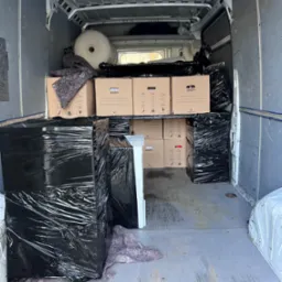 Wnętrze busa transportowego wypełnione kartonami i meblami owiniętymi folią stretch, przygotowanymi do przewozu. Widoczne zabezpieczenia i opakowania.