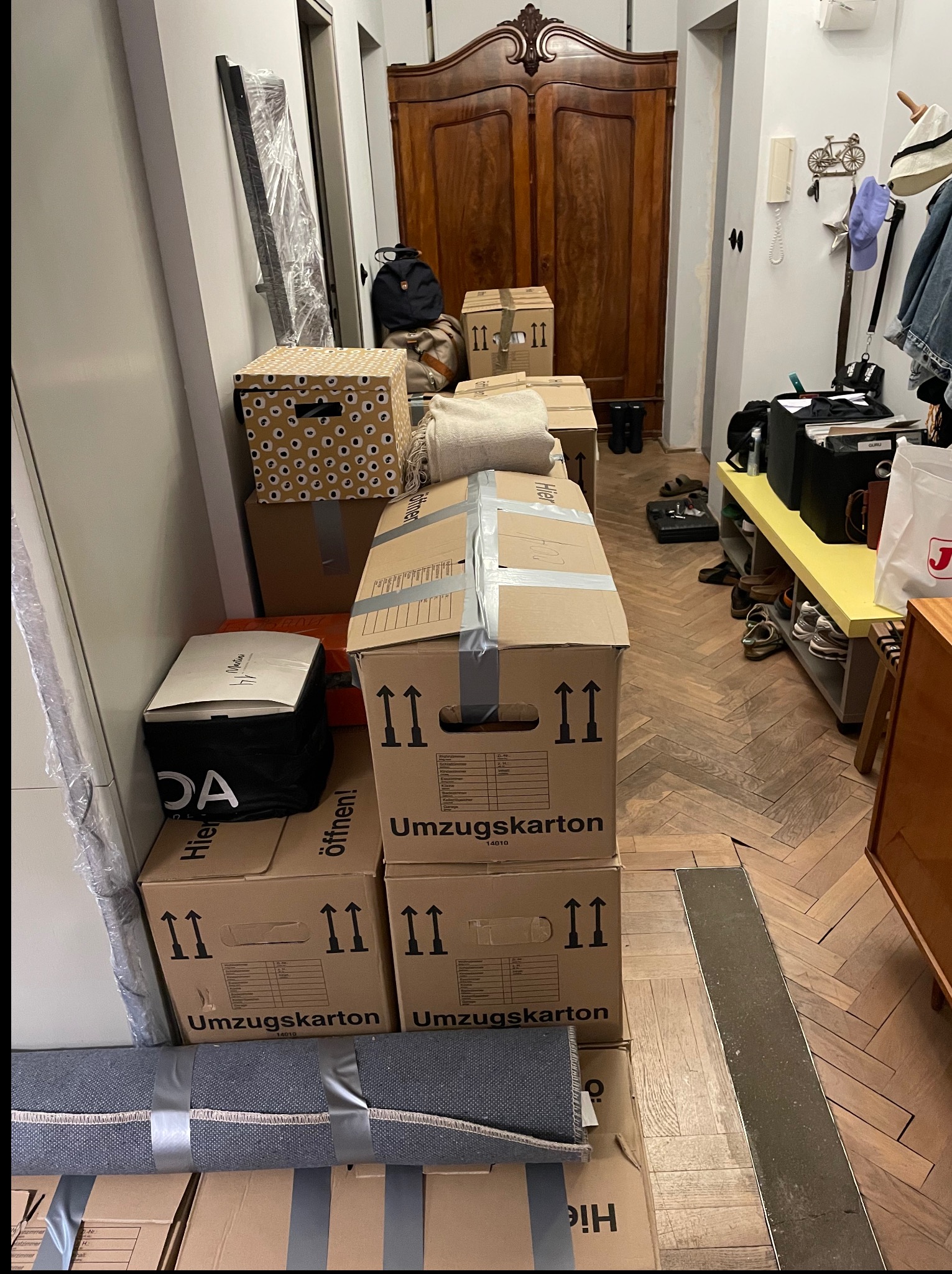 Korytarz mieszkania wypełniony kartonami do przeprowadzki, z widocznym parkietem w jodełkę i starą szafą. Kartony z napisem 'Umzugskarton' i strzałkami do góry.