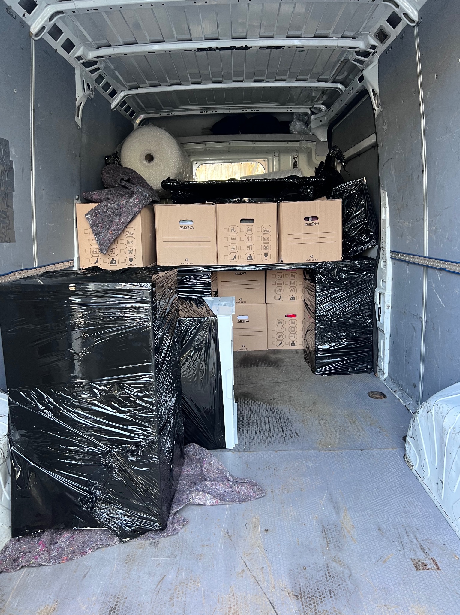 Wnętrze busa transportowego wypełnione kartonami i meblami owiniętymi folią stretch, przygotowanymi do przewozu. Widoczne zabezpieczenia i opakowania.