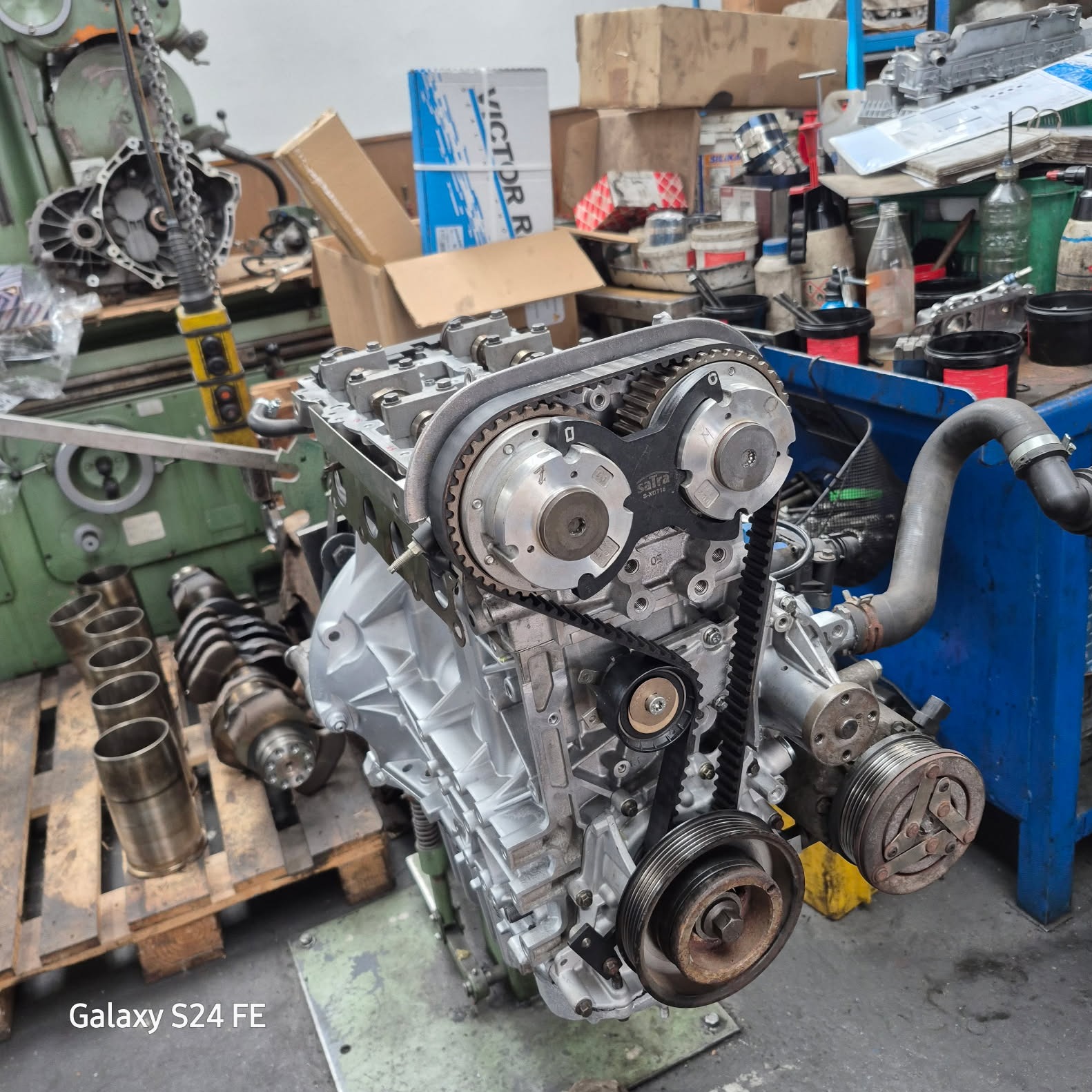 Szczegółowe ujęcie odnowionego silnika samochodowego z nowym paskiem rozrządu na tle warsztatu z narzędziami i częściami, ukazujące precyzję i dbałość o detale.