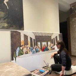 Sztuco-Art - Artystka maluje replikę 'Ostatniej Wieczerzy' na płótnie w jasnym wnętrzu. Widoczne pędzle, farby i inne obrazy w tle. Praca w toku.