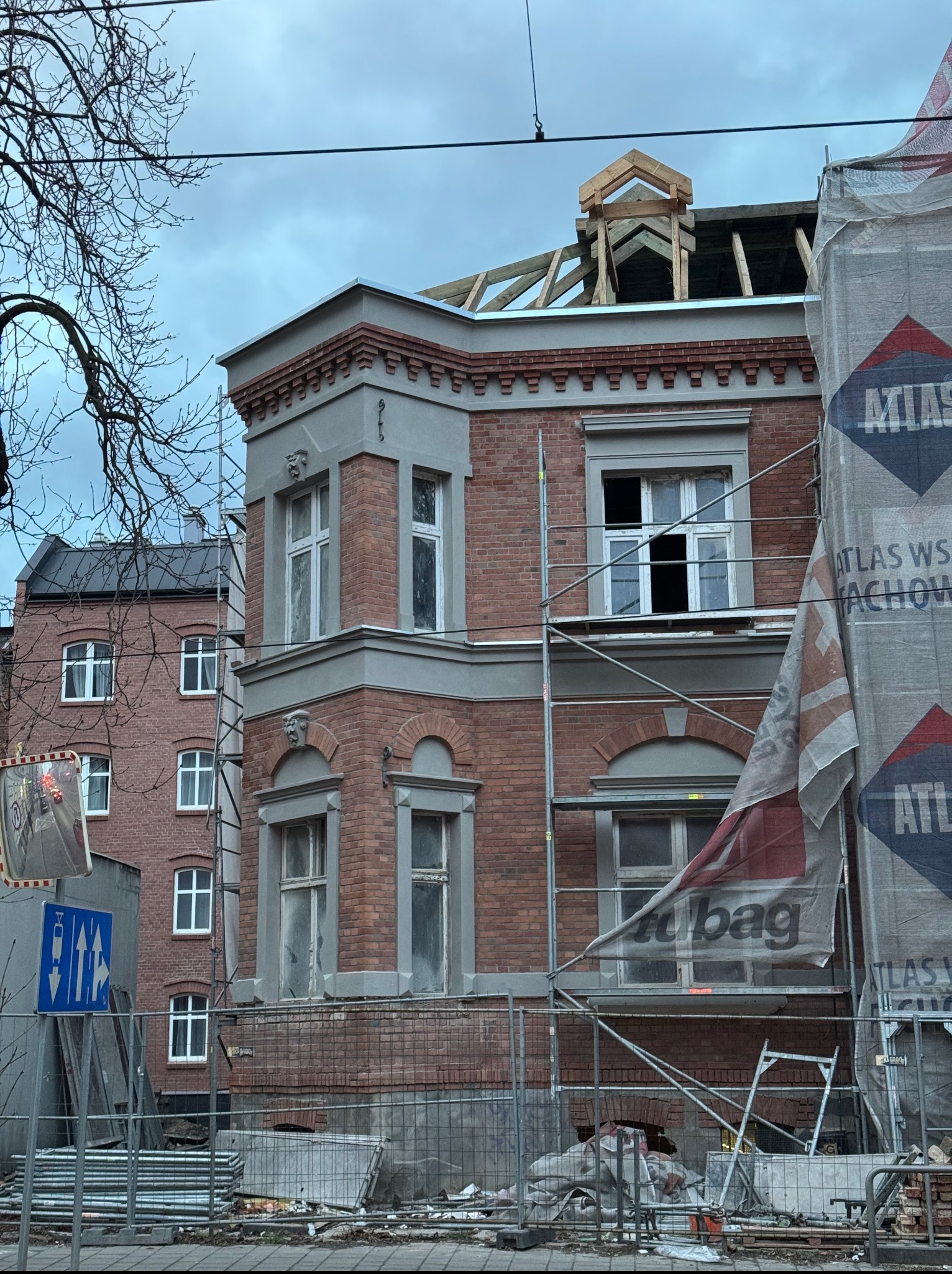 Renowacja ceglanej kamienicy z rusztowaniem i odsłoniętą konstrukcją dachu, widok z dołu na budynek w trakcie prac, z fragmentem sąsiedniej zabudowy.