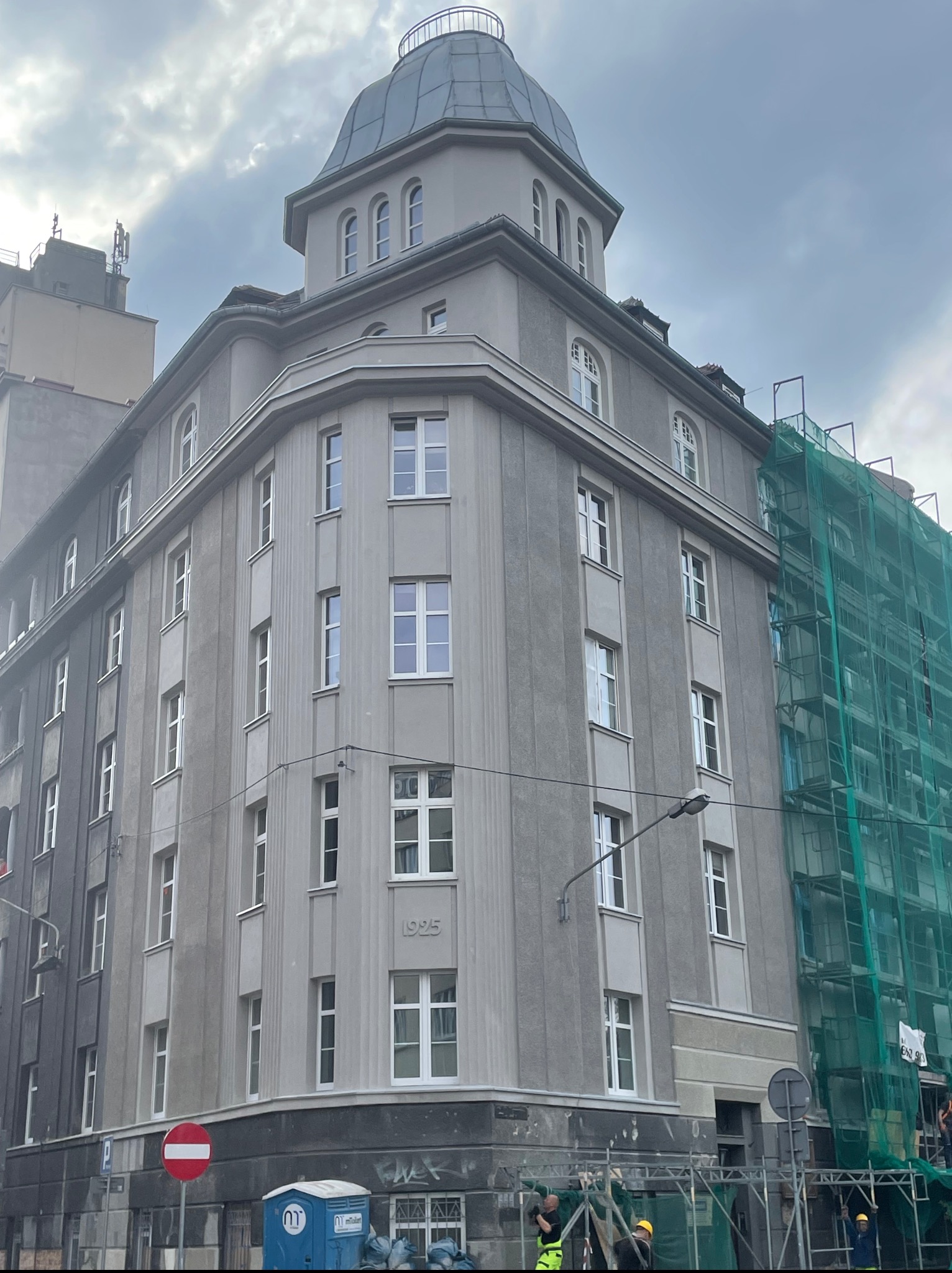 Szara kamienica z 1925 roku z kopułą i rusztowaniem, częściowo zakryta siatką ochronną, sugerująca renowację elewacji. Widoczny znak drogowy i przenośna toaleta.