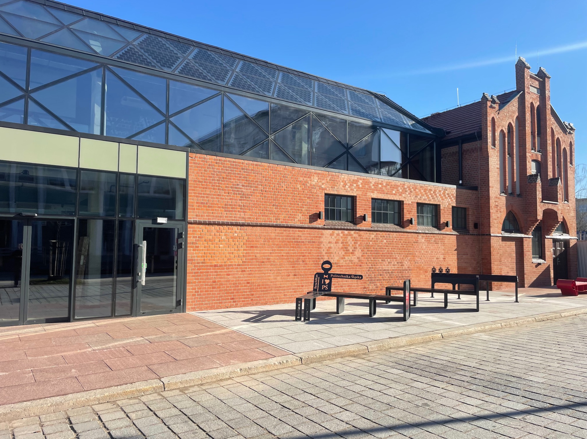 Budynek z czerwonej cegły z nowoczesną szklaną konstrukcją. Elementy zabytkowe i nowoczesne. Widoczne ławki i chodnik z kostki.