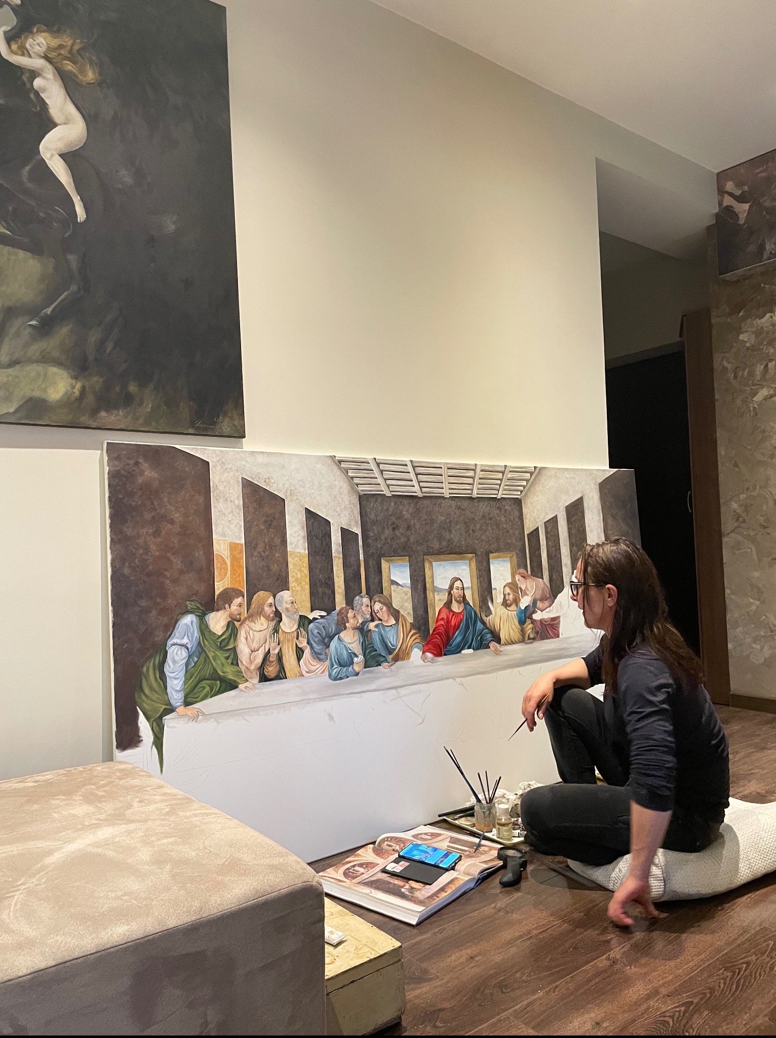 Artystka maluje replikę 'Ostatniej Wieczerzy' na płótnie w jasnym wnętrzu. Widoczne pędzle, farby i inne obrazy w tle. Praca w toku.