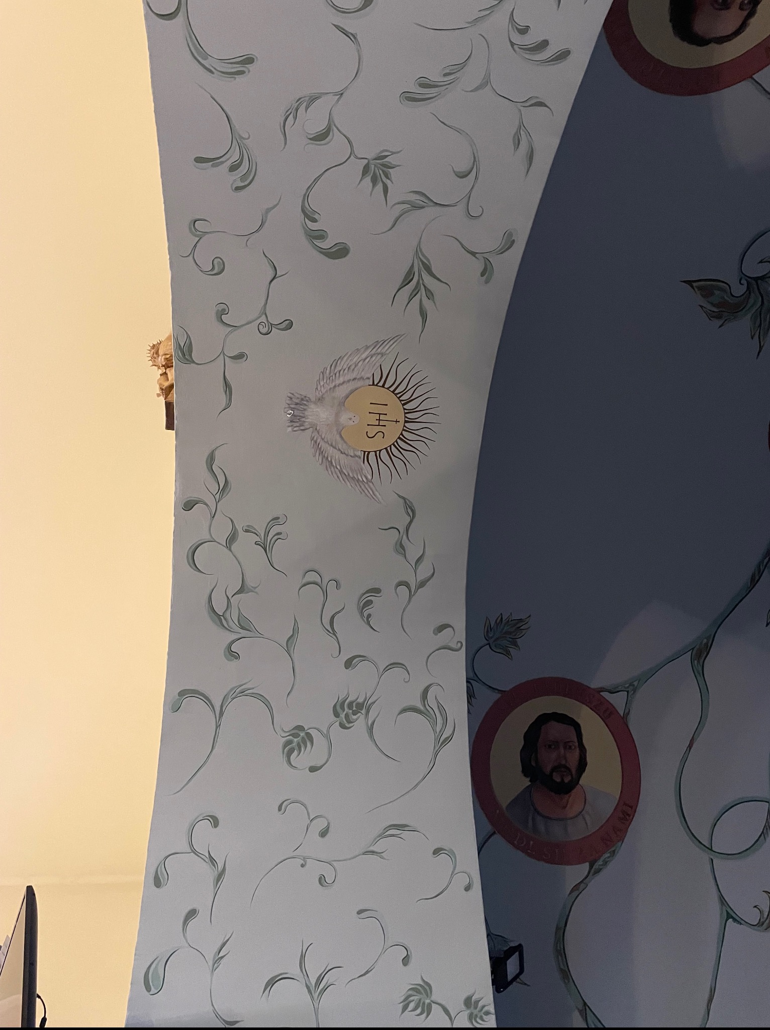 Malowidło na sklepieniu z motywami roślinnymi, wizerunkami świętych i symbolami religijnymi, wykonane w jasnej kolorystyce. Detal z bliska.