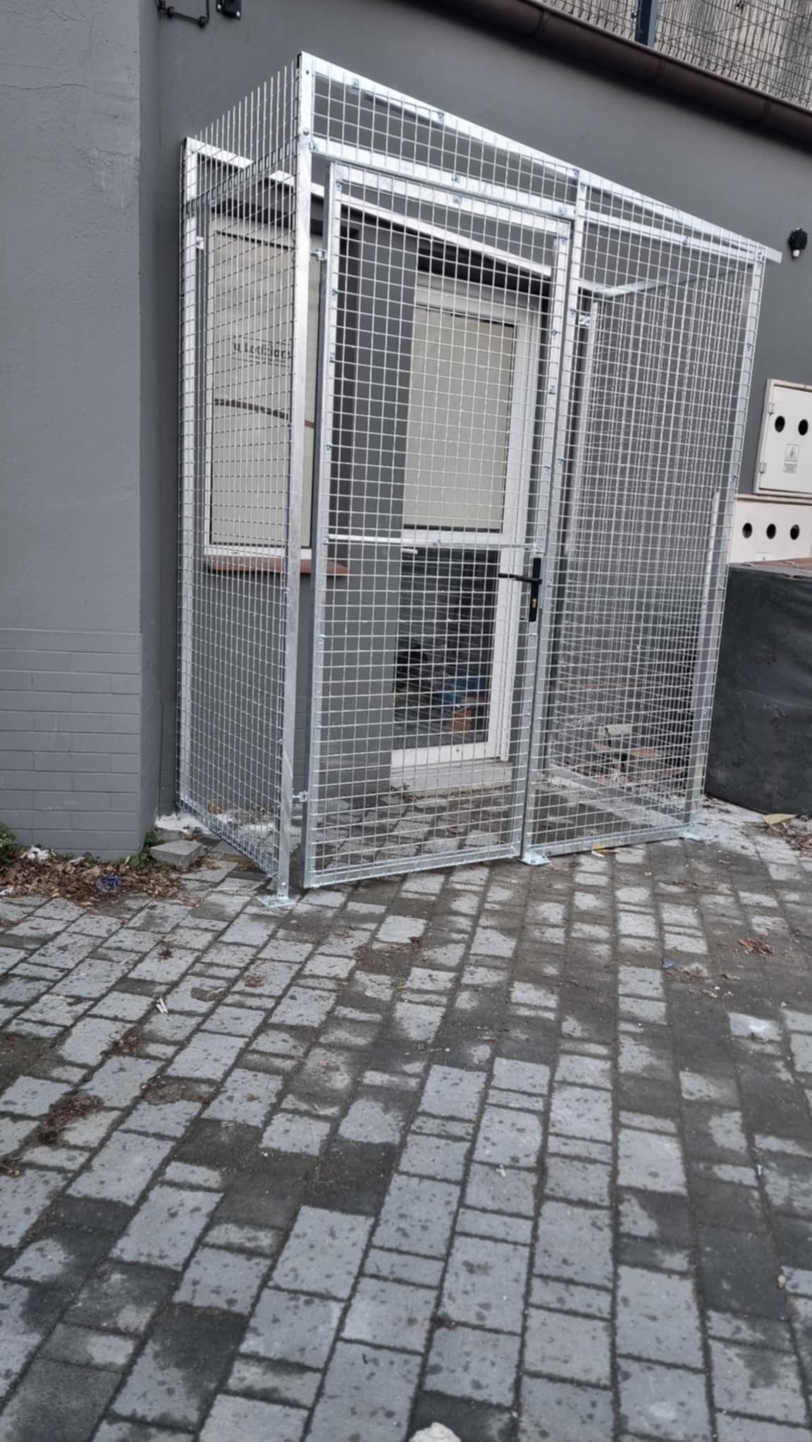 Metalowa klatka ochronna z siatki, zabezpieczająca drzwi wejściowe do budynku. Widoczne spawane łączenia i szara elewacja.