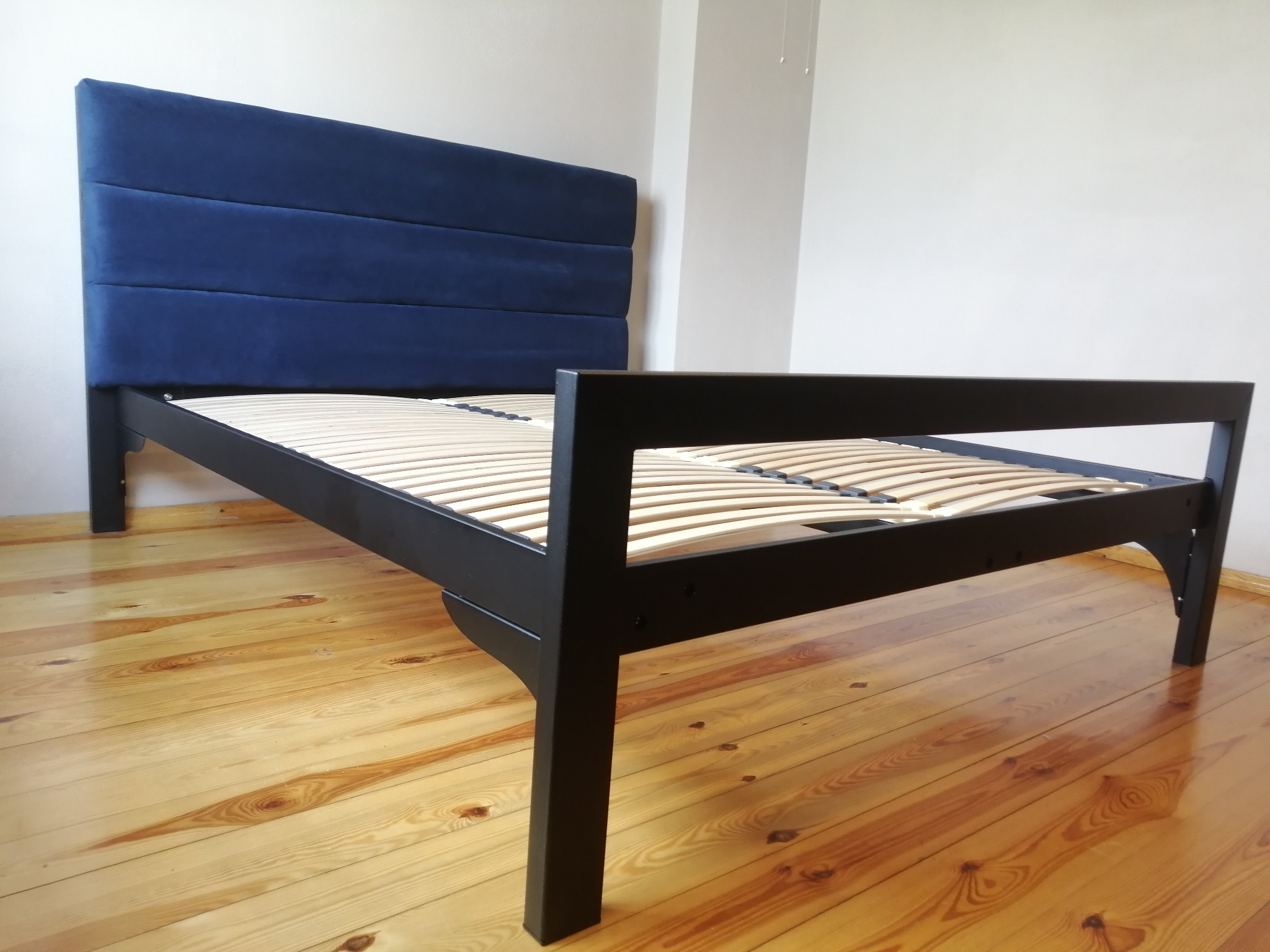 Czarne, metalowe łóżko z drewnianym stelażem i tapicerowanym, granatowym wezgłowiem na drewnianej podłodze. Minimalistyczny design, proste linie, widoczne detale łączeń.