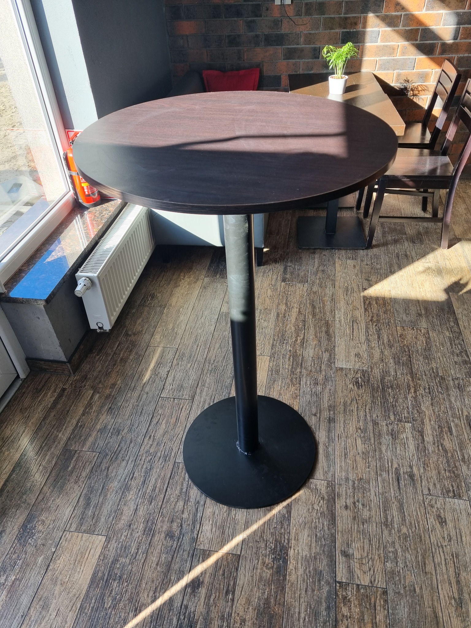 Stół barowy z ciemnym blatem i czarną nogą na tle drewnopodobnych płytek podłogowych, z widocznym fragmentem okna i grzejnika.