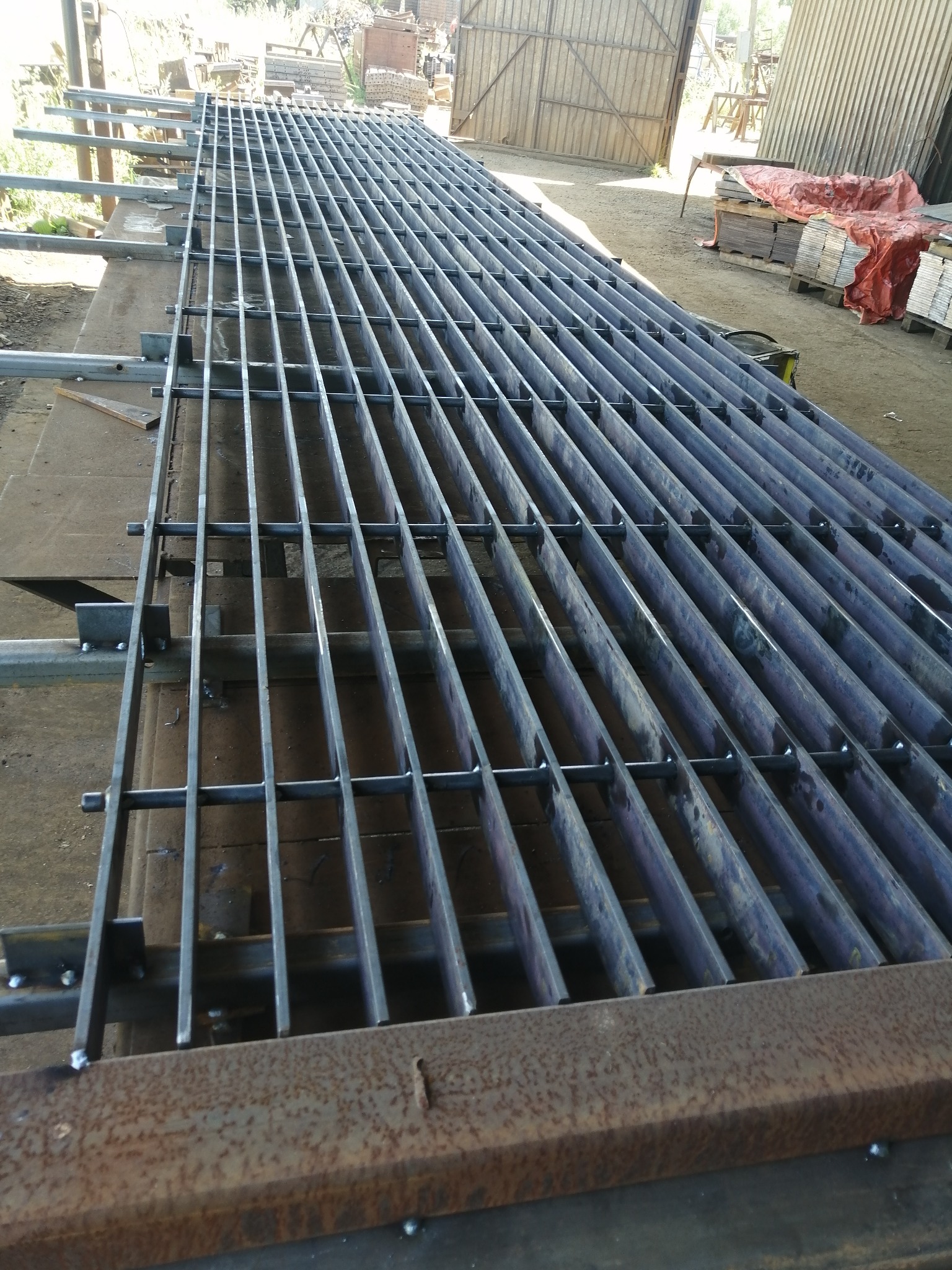 Konstrukcja kraty metalowej z rur i płaskowników, spawana na stole warsztatowym. Widoczne ślady spawania, surowy metal, warsztatowy klimat.