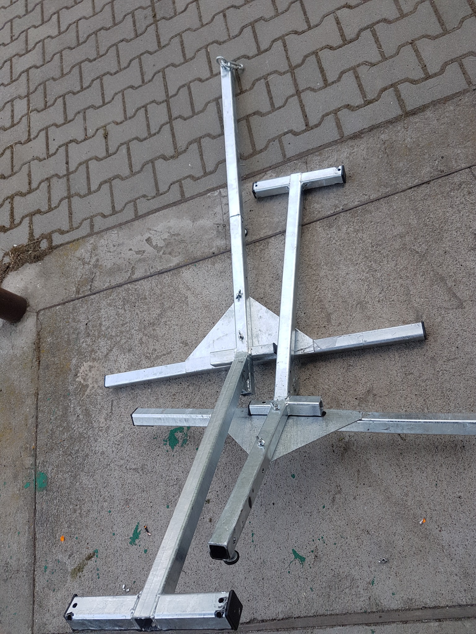 Dwa srebrne, metalowe stojaki o trójkątnych podstawach i regulowanej wysokości, leżące na szarym chodniku z kostki. Widoczne ślady użytkowania.