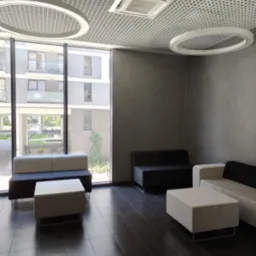 Nowoczesne wnętrze biurowe z sofami i stolikami w odcieniach szarości, widok na zewnątrz przez duże okno, oświetlenie sufitowe w kształcie okręgów.