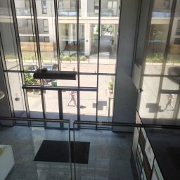 TAXENDO SPÓŁKA Z OGRANICZONĄ ODPOWIEDZIALNOŚCIĄ - Wnętrze nowoczesnego biurowca z widokiem na dziedziniec przez szklaną fasadę. Osoba przechodzi przez wejście. Marmurowa podłoga i panel reklamowy.