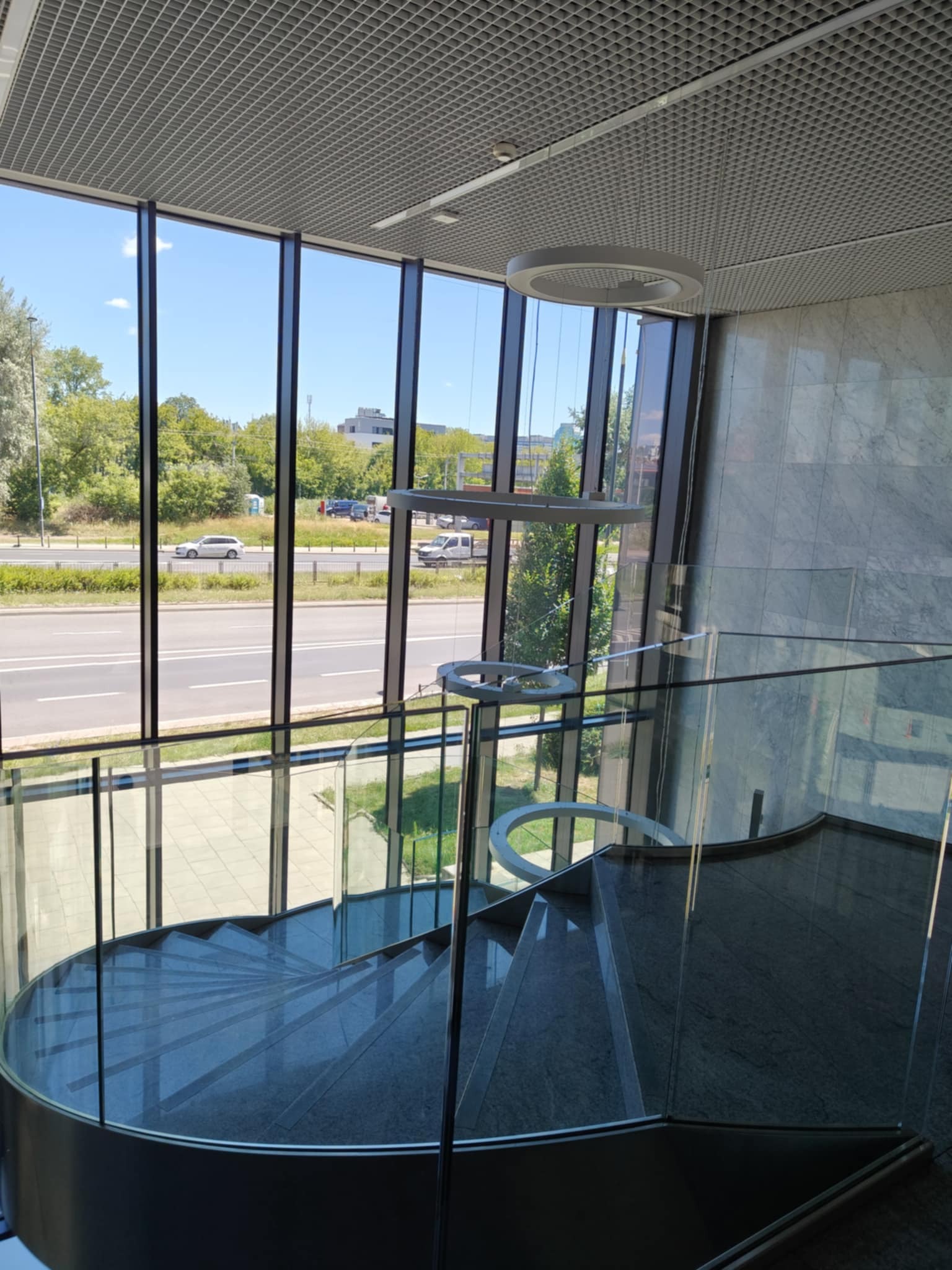 Wnętrze biurowca: marmurowe schody spiralne z przeszkloną balustradą, widok na ulicę przez wysokie okna, nowoczesna lampa wisząca. Jasne, przestronne wnętrze.