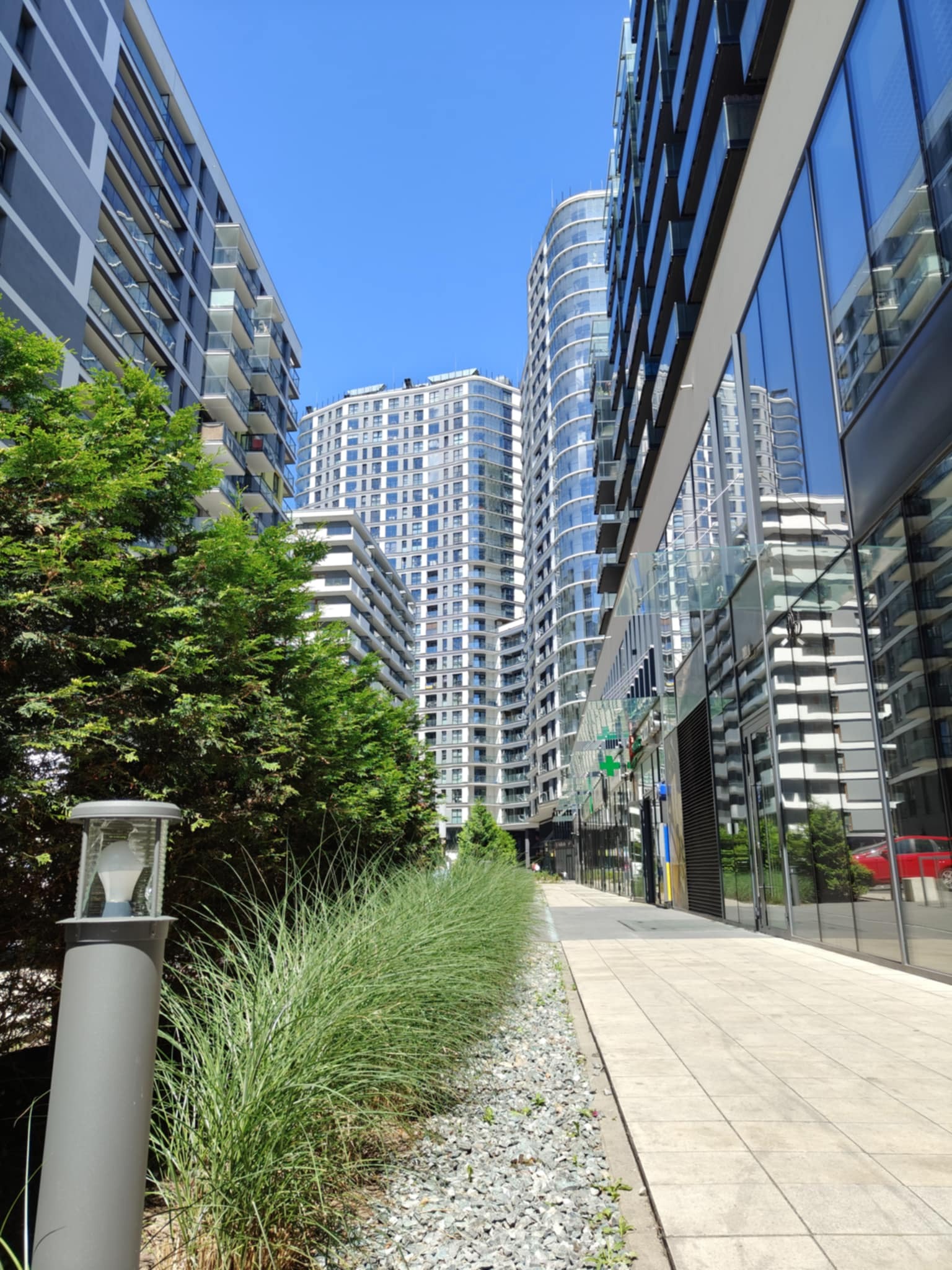 Nowoczesna architektura miejska: chodnik z kostki, zieleń i wysokie budynki z przeszklonymi fasadami w słoneczny dzień. Widok z dołu na błękitne niebo.