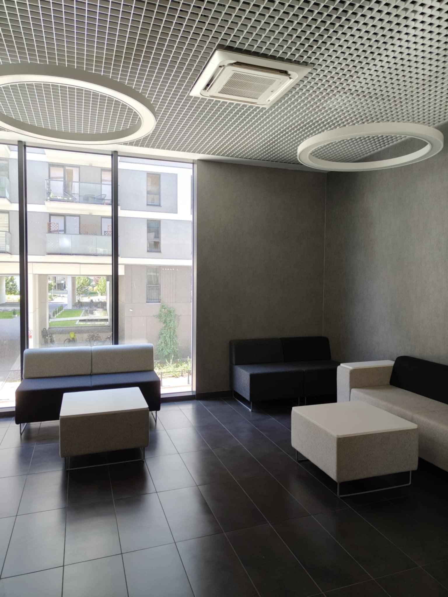Nowoczesne wnętrze biurowe z sofami i stolikami w odcieniach szarości, widok na zewnątrz przez duże okno, oświetlenie sufitowe w kształcie okręgów.