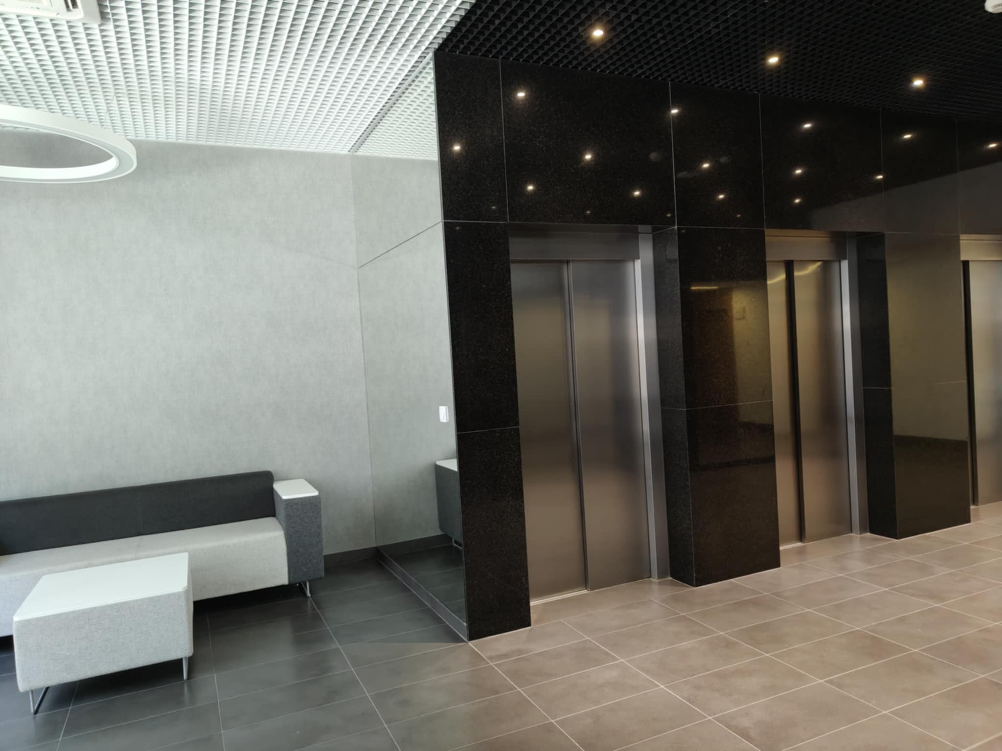 Nowoczesne wnętrze biurowe z sofą, stolikiem i windami. Czarno-biała kolorystyka, oświetlenie sufitowe. Minimalistyczny design.