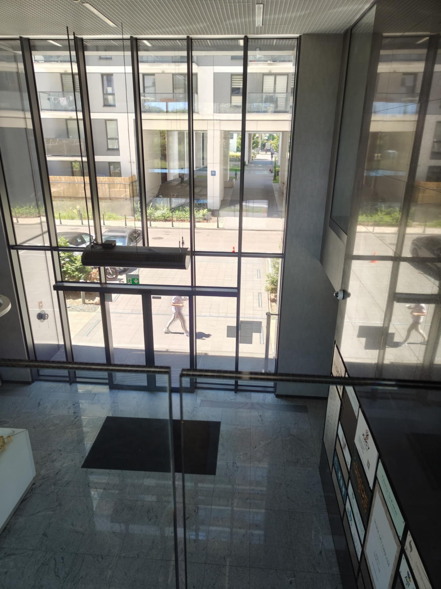 Wnętrze nowoczesnego biurowca z widokiem na dziedziniec przez szklaną fasadę. Osoba przechodzi przez wejście. Marmurowa podłoga i panel reklamowy.