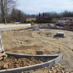 Plac budowy z wytyczonym okręgiem z obrzeży betonowych, przygotowany podłoże, mini koparka i paleta z pojemnikiem na wodę. W tle budynek i zalesiony teren.