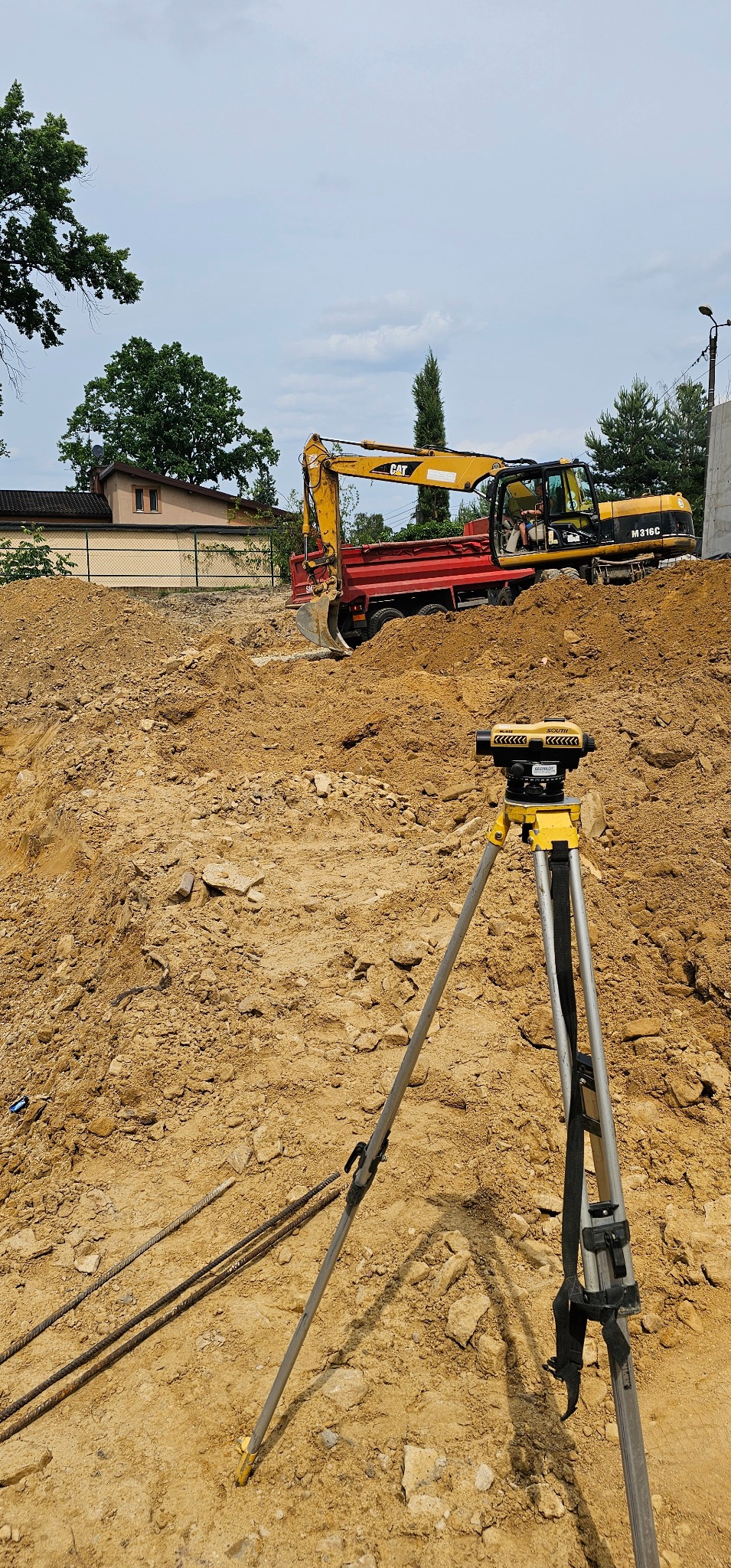 Żółty niwelator optyczny na statywie na placu budowy z wykopem i koparką w tle, w słoneczny dzień.