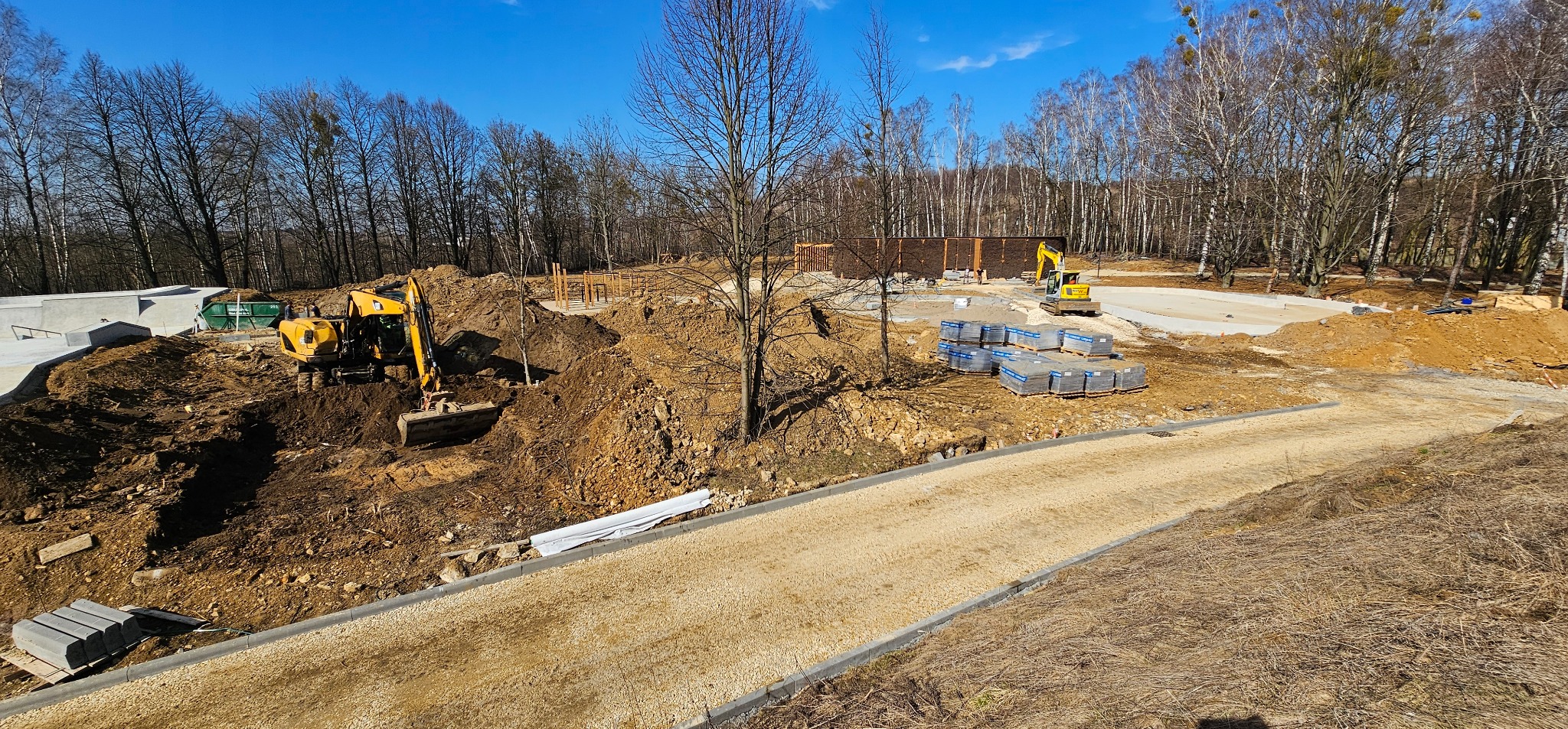 Krajobraz placu budowy z dwoma żółtymi koparkami, paletami kostki i fragmentami ułożonej nawierzchni w słoneczny dzień. W tle drzewa i konstrukcja.
