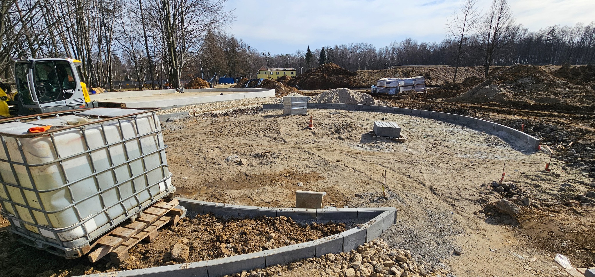 Plac budowy z wytyczonym okręgiem z obrzeży betonowych, przygotowany podłoże, mini koparka i paleta z pojemnikiem na wodę. W tle budynek i zalesiony teren.