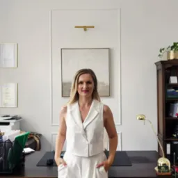 Portret adwokatki w biurze: blond kobieta w białym kostiumie na tle minimalistycznego wnętrza z regałem książek i dokumentami.