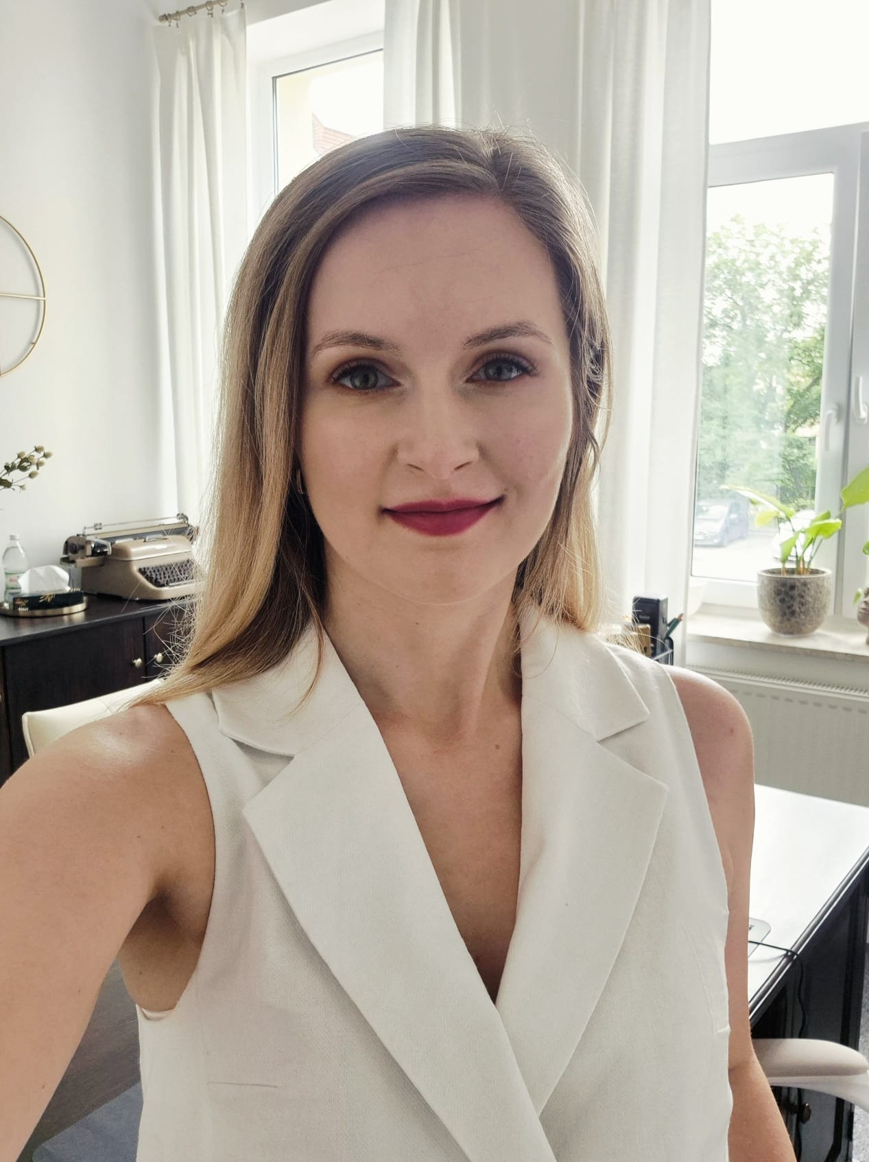 Portret kobiety w białej kamizelce na tle jasnego biura z maszyną do pisania i roślinami, sugerujący profesjonalizm adwokata.