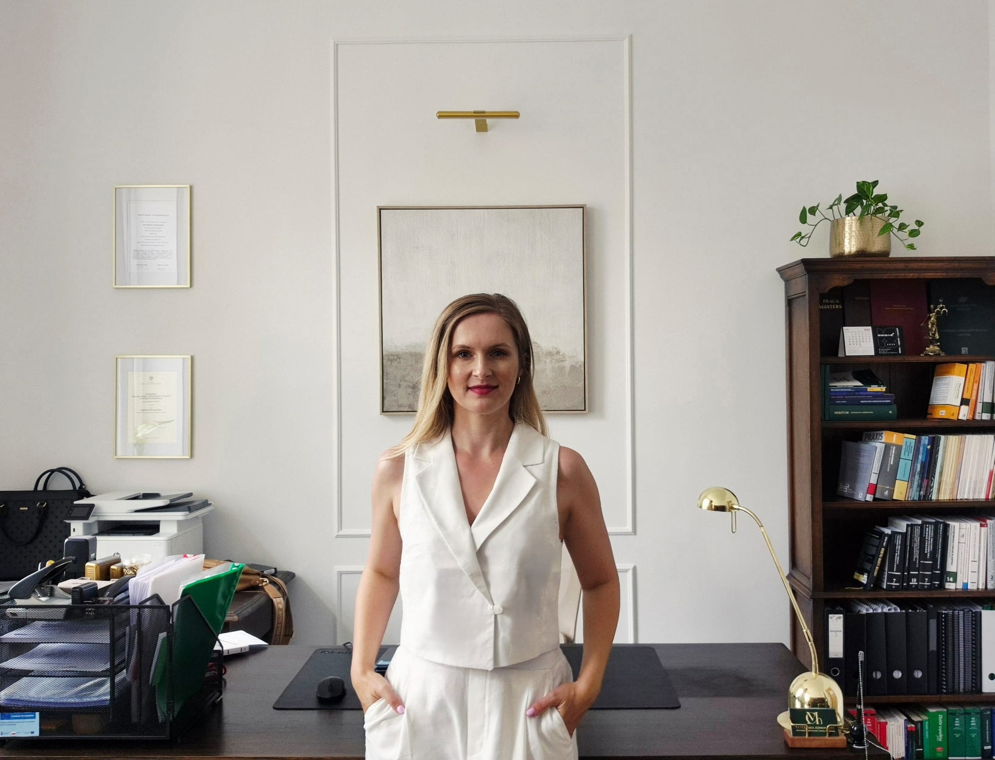 Portret adwokatki w biurze: blond kobieta w białym kostiumie na tle minimalistycznego wnętrza z regałem książek i dokumentami.