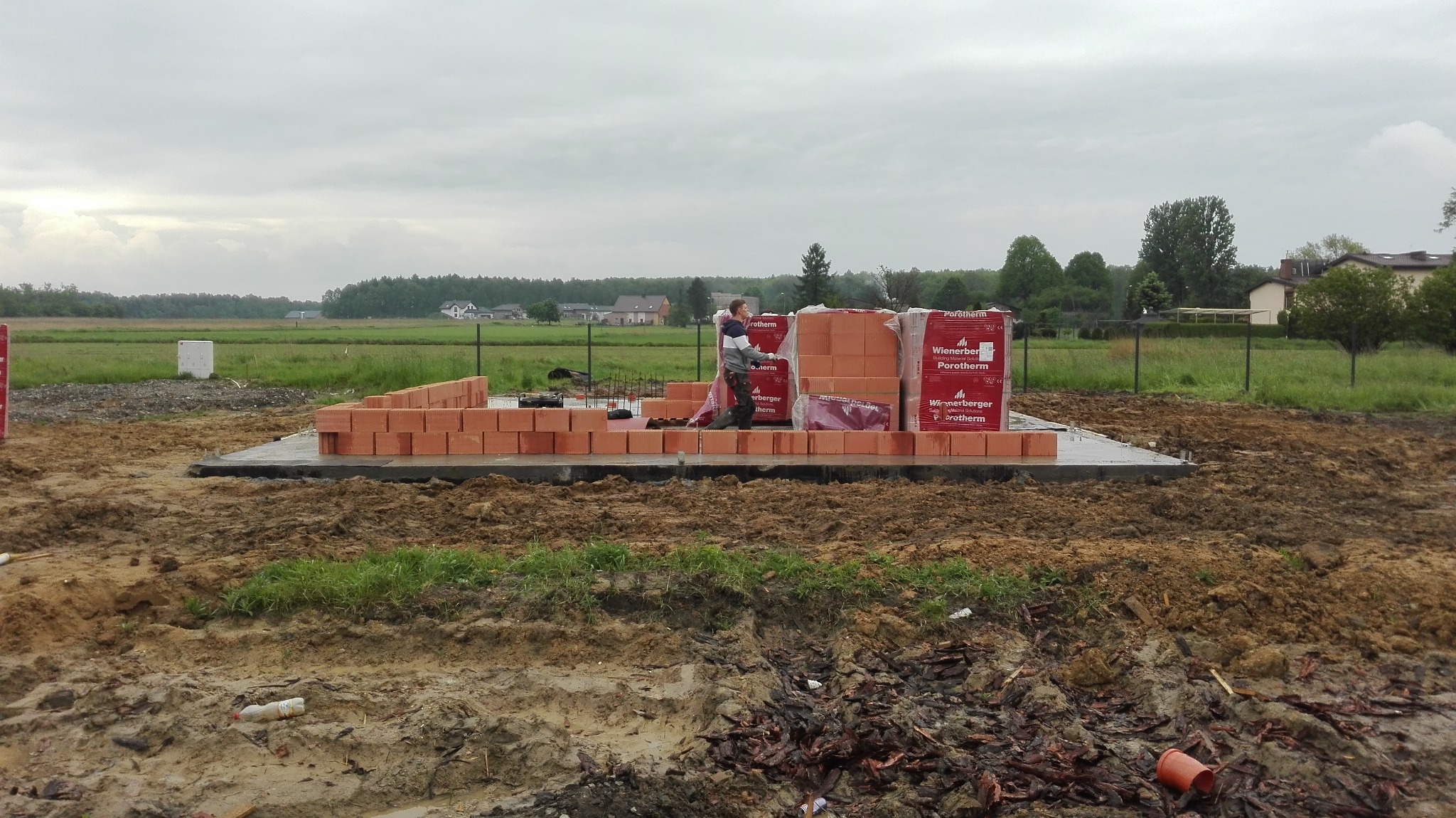 Budowa domu w toku: murarz układa pierwszą warstwę cegieł Porotherm na fundamencie. W tle krajobraz wiejski i pochmurne niebo. Widoczne palety z cegłami.
