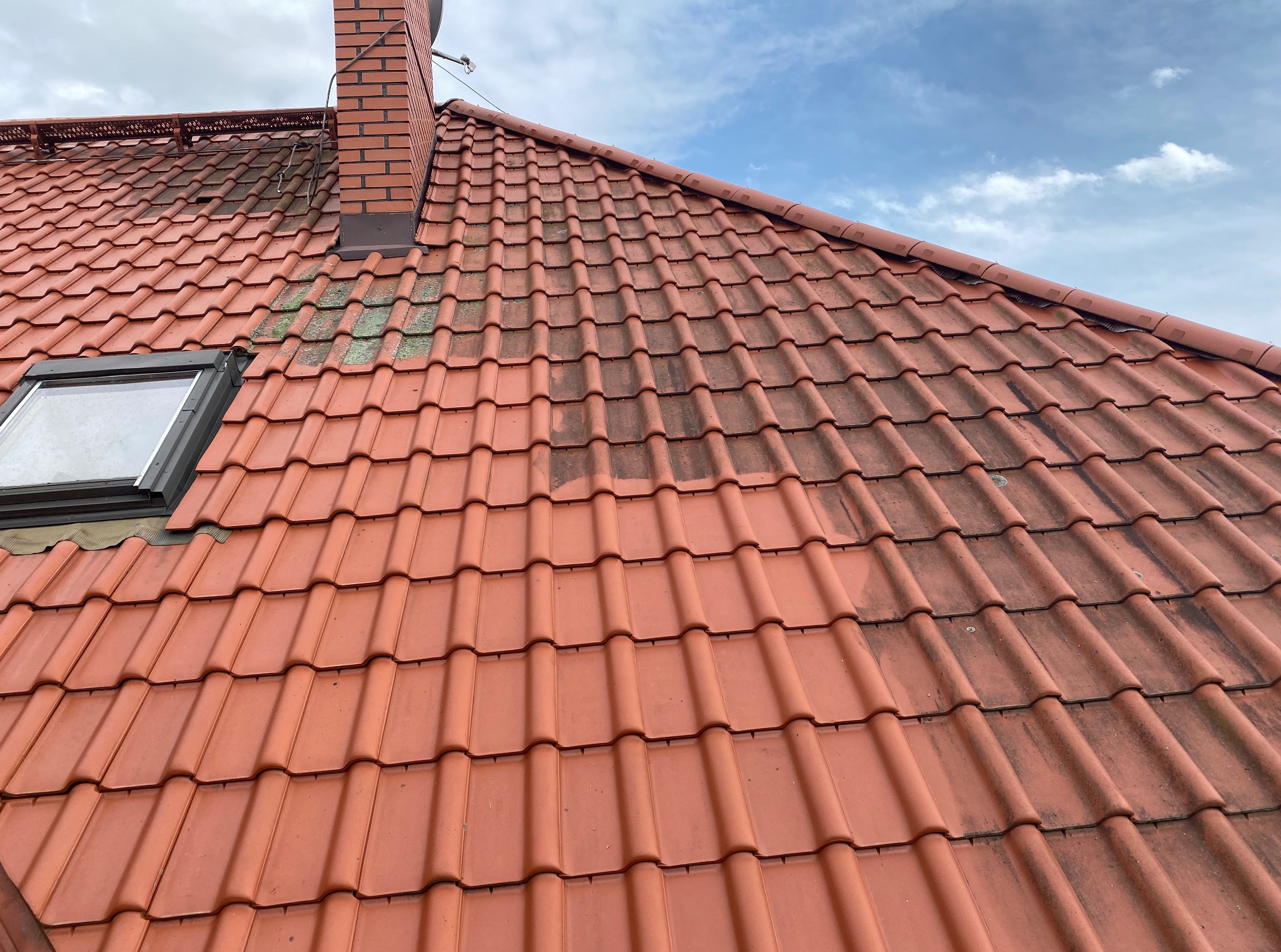 Częściowo wyczyszczony, czerwony dach z dachówki ceramicznej, widoczny komin z cegły i okno dachowe. Połowa dachu czysta, druga pokryta zabrudzeniami i mchem.