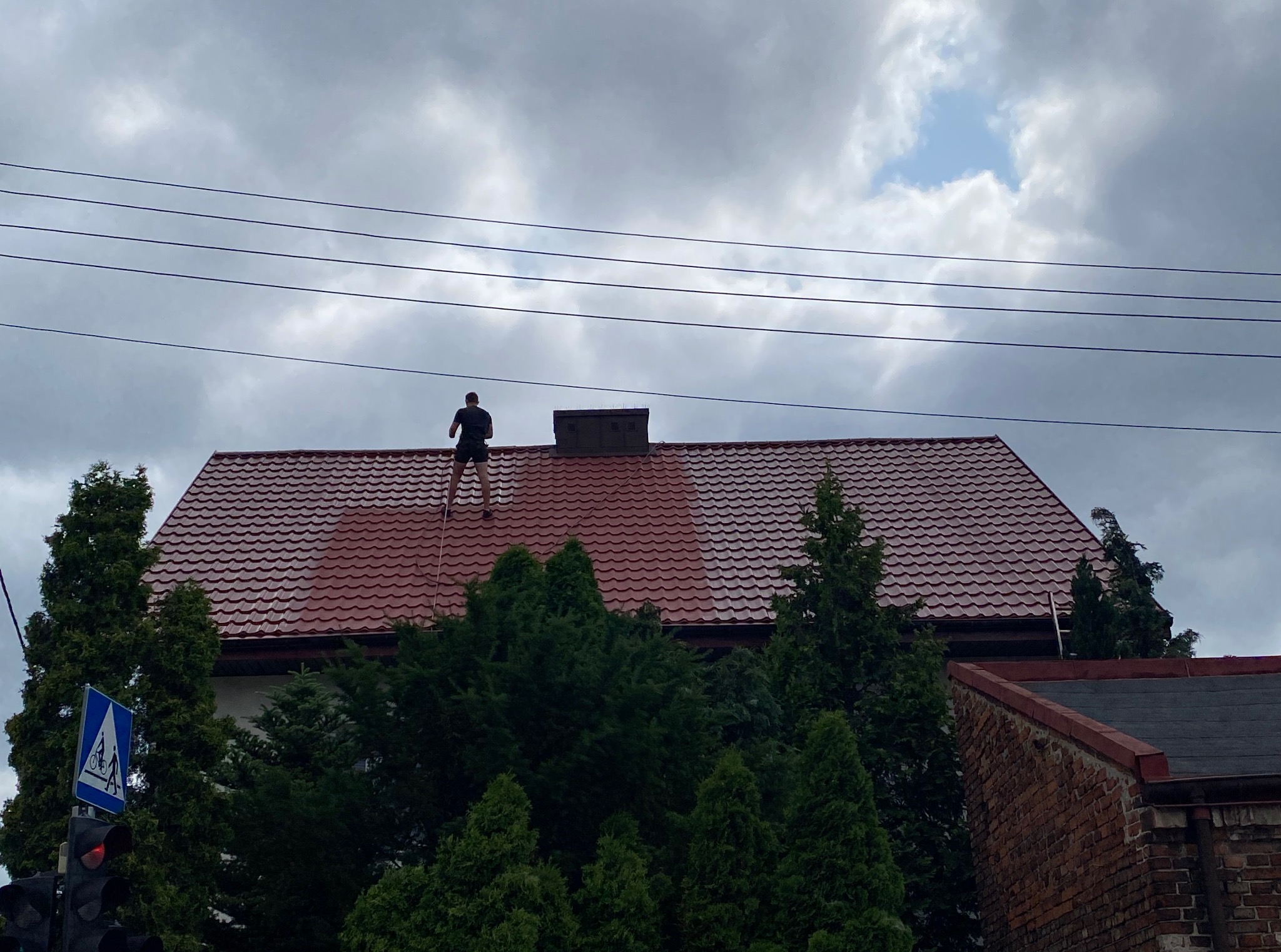 Mężczyzna na dachu z czerwonej dachówki, w trakcie malowania. Widoczne świeżo pomalowane fragmenty dachu i drabina oparta o dach. W tle pochmurne niebo i zieleń drzew.