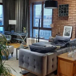 Loftowe wnętrze z ceglaną ścianą, szarą sofą i drewnianą podłogą. Pies śpi pod sofą. Widoczne okno z żaluzjami i dekoracje.