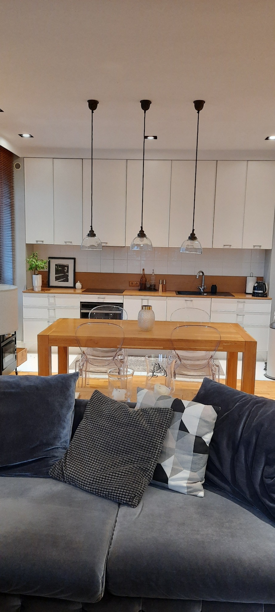 Nowoczesna kuchnia z drewnianym stołem i przezroczystymi krzesłami, widok z salonu z szarą sofą i dekoracyjnymi poduszkami w stonowanych barwach.