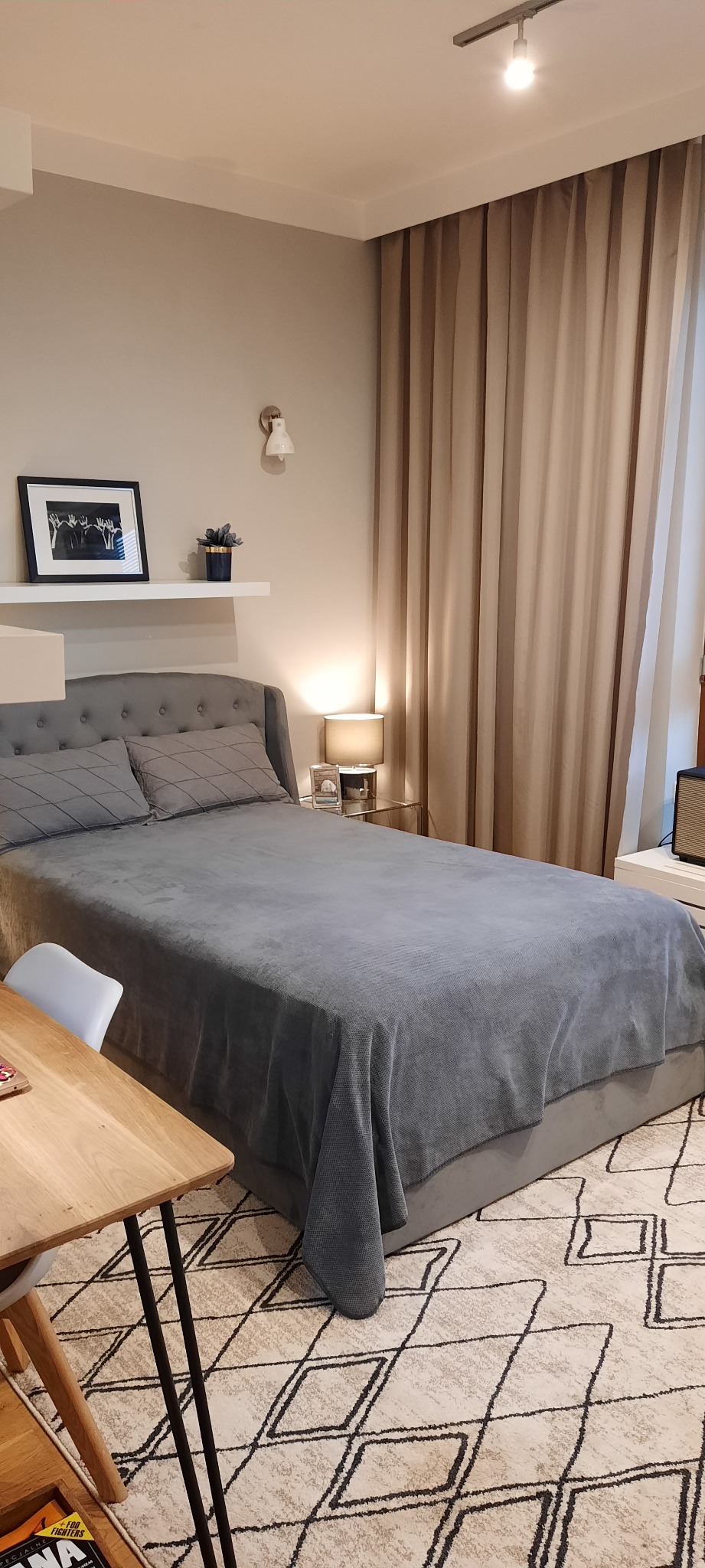 Sypialnia w odcieniach szarości i beżu: łóżko z szarym zagłówkiem i narzutą, drewniany stolik, wzorzysty dywan. Nad łóżkiem półka z dekoracjami i lampka.