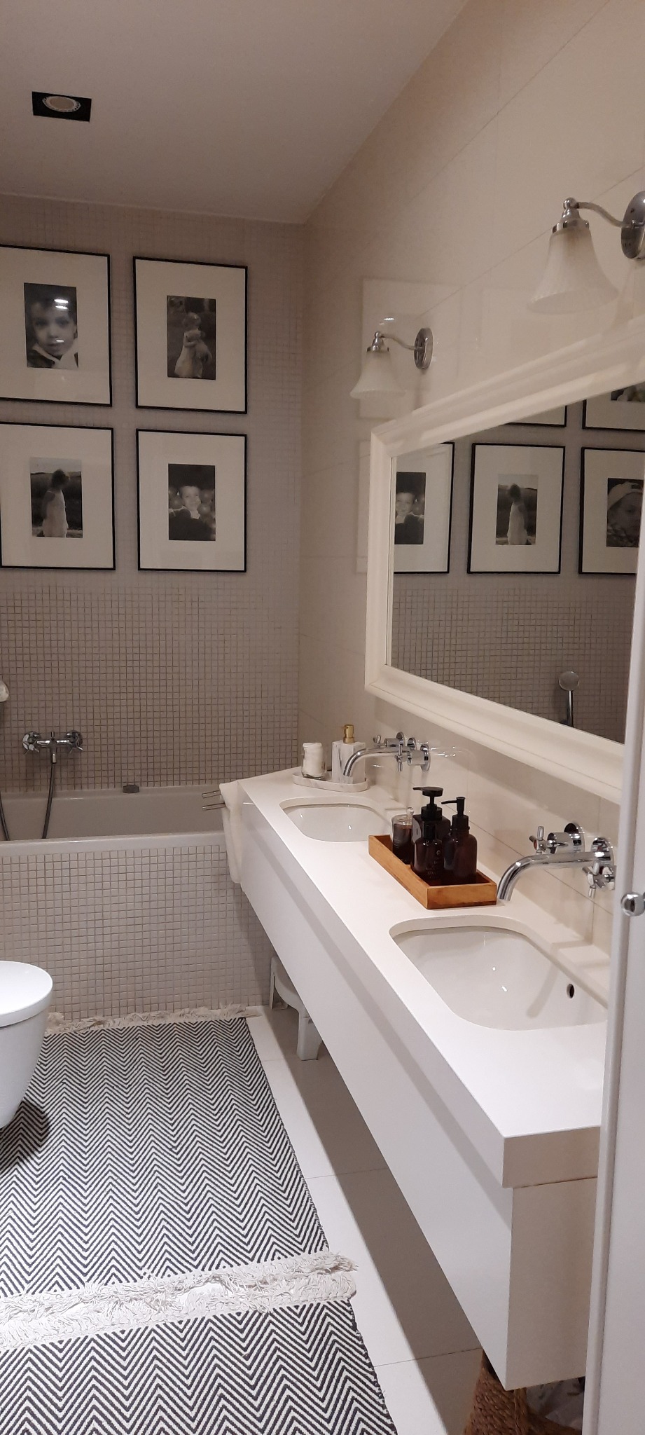 Nowoczesna łazienka z dwoma umywalkami, lustrem i galerią czarno-białych zdjęć w ramkach na ścianie. Wzornisty dywanik na podłodze.